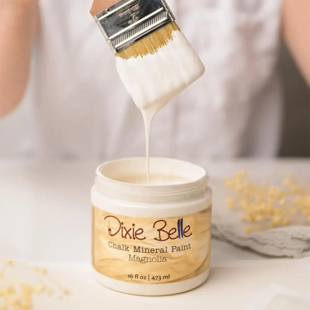 A paint brush with dripping paint is shown above an open container of Dixie Belle's Magnolia Chalk Mineral Paint.