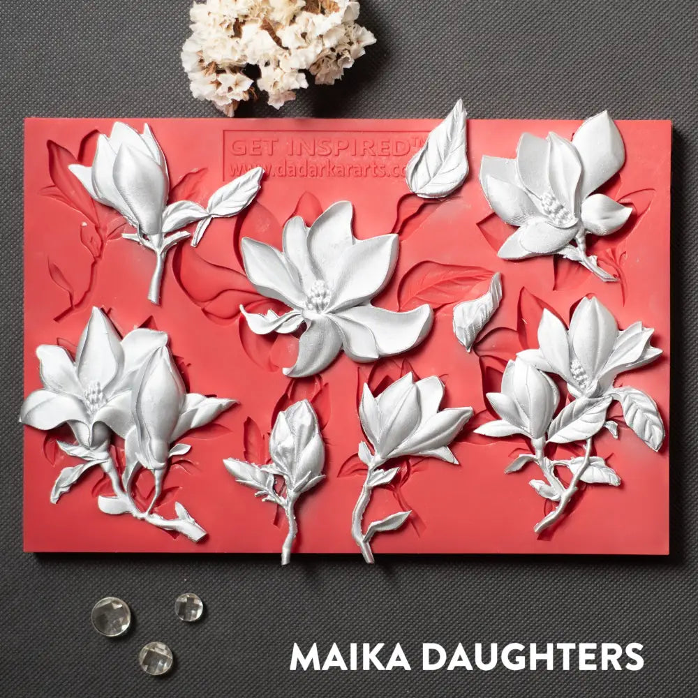 A red silicone mold and silver colored castings of 8 magnolia flower blooms and 2 leaves are against a dark grey material background. 