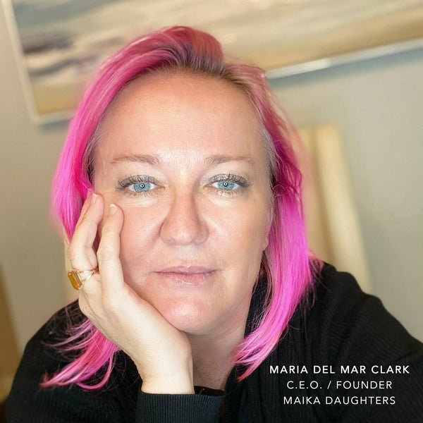 A woman in her late 40's staring into the camera with her face on her hand, and bright pink hair. On the bottom right hand corner it says: MARIA DEL MAR CLARK, CEO / FOUNDER, MAIKA DAUGHTERS