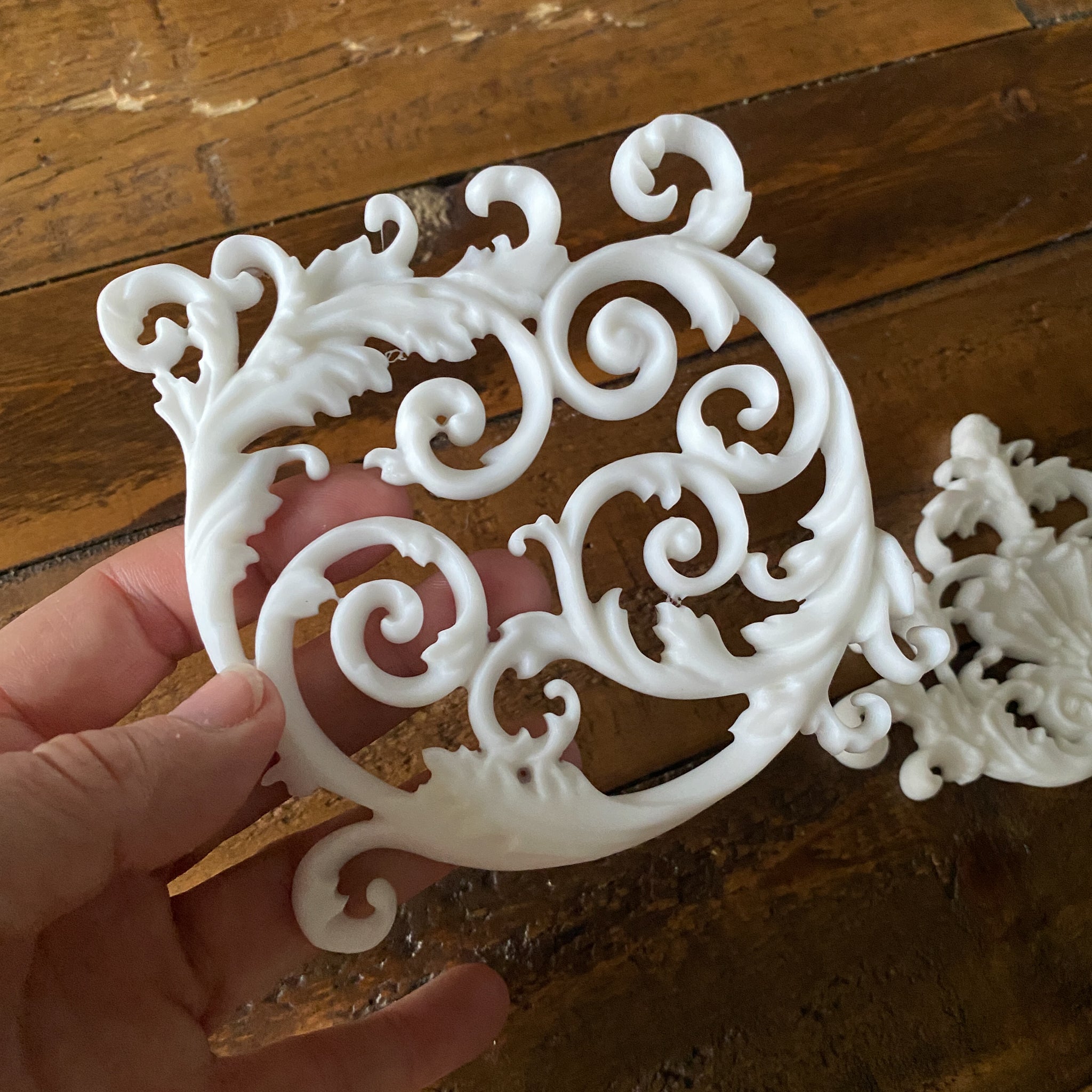 Two white resin castings of ornate scroll center accent pieces are against a wood background. A hand is shown holding one of the castings.