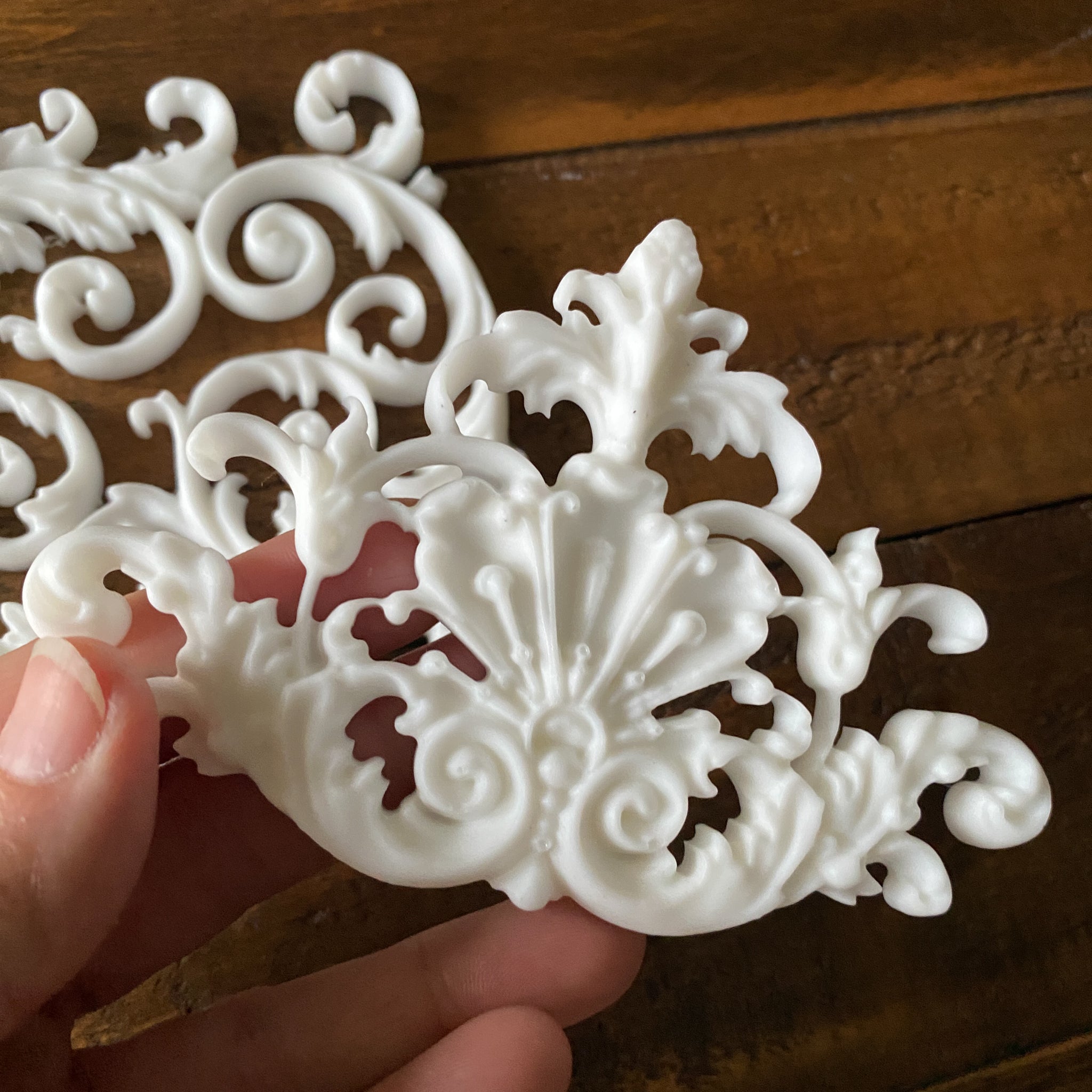 Two white resin castings of ornate scroll center accent pieces are against a wood background. A hand is shown holding one of the castings.