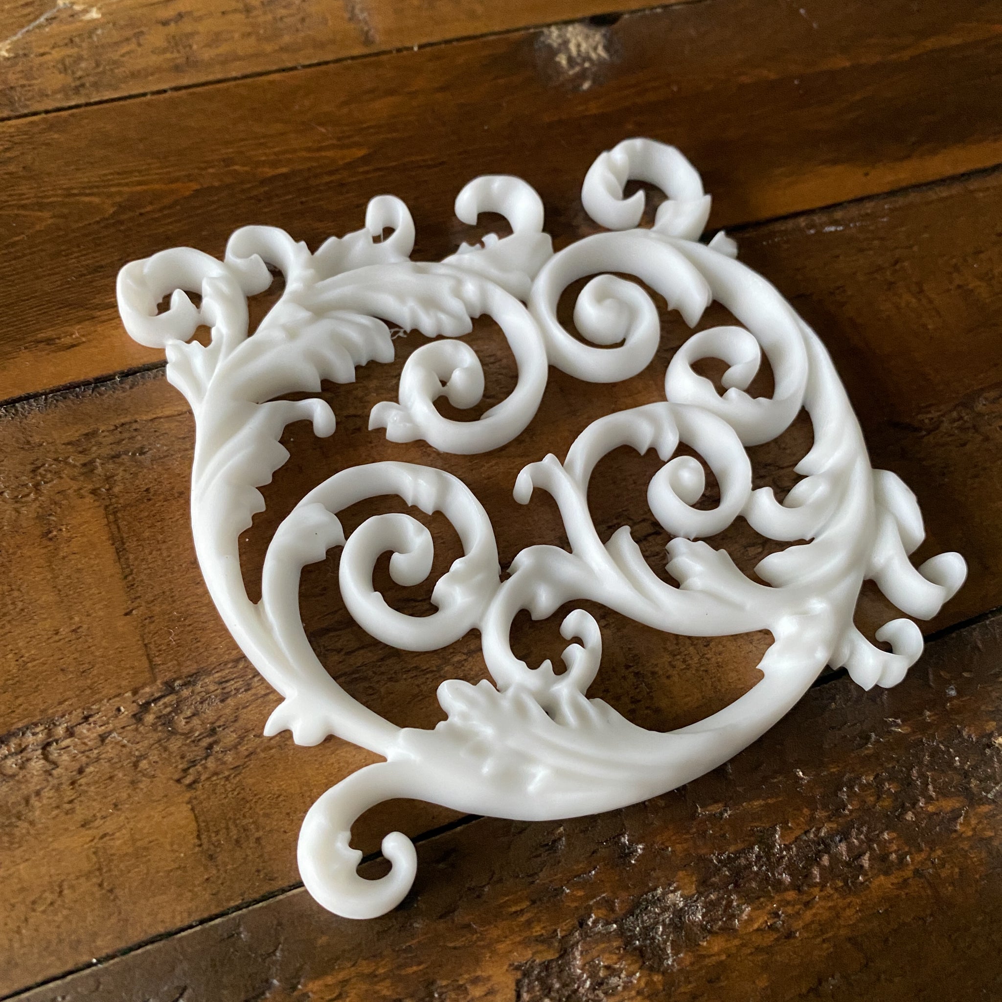 A white resin castings of an ornate scroll center accent pieces is against a wood background.