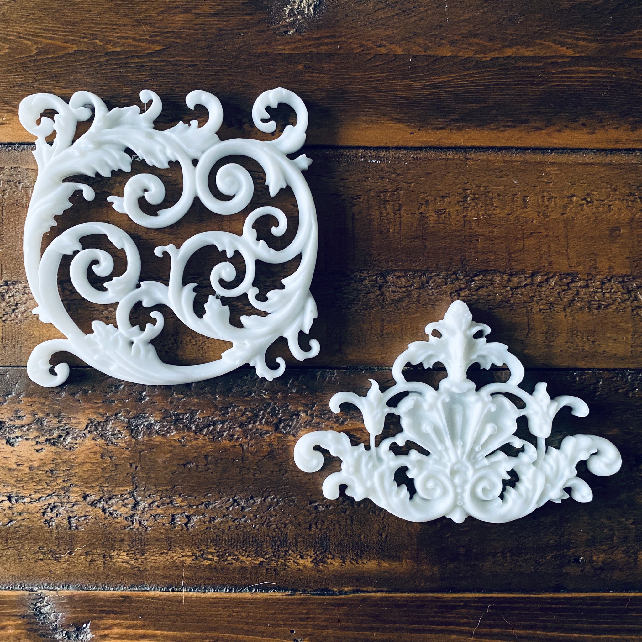 Two white resin castings created from LaBlanche's Ornament Claudette silicone mold of ornate scroll center accent pieces are against a wood background.