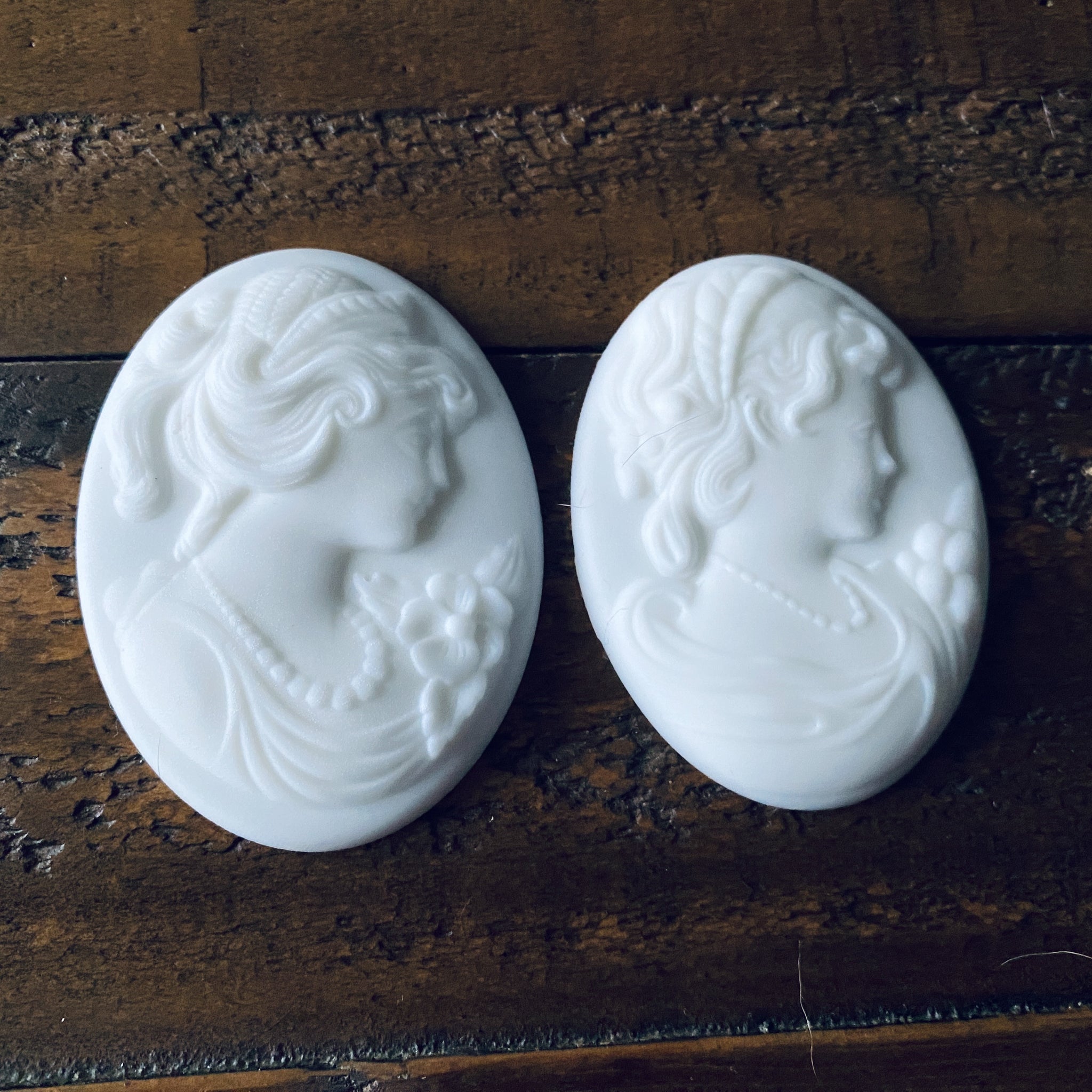 A close-up of 2 white resin cameo pendants created from LaBlanche's Vintage Cameo silicone mold are against a wood background.