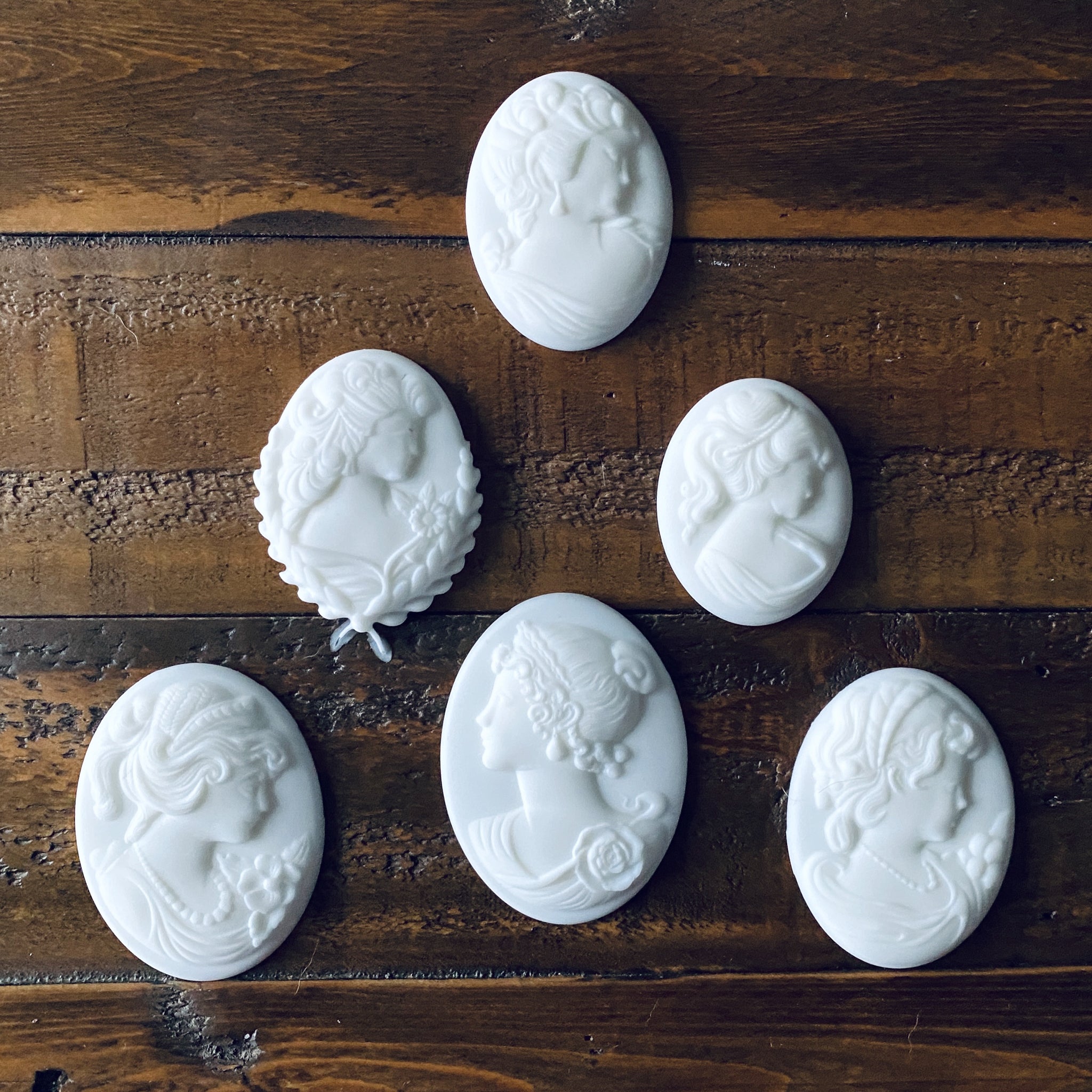 Six white resin cameo pendants created from LaBlanche's Vintage Cameo silicone mold are against a wood background.