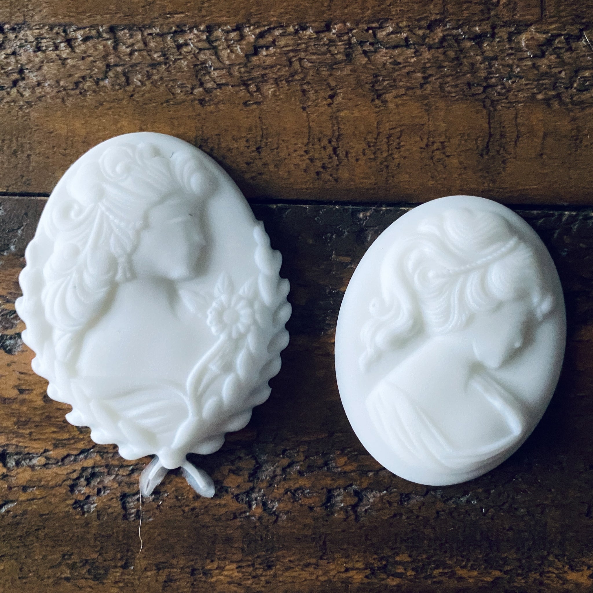 A close-up of 2 white resin cameo pendants created from LaBlanche's Vintage Cameo silicone mold are against a wood background.