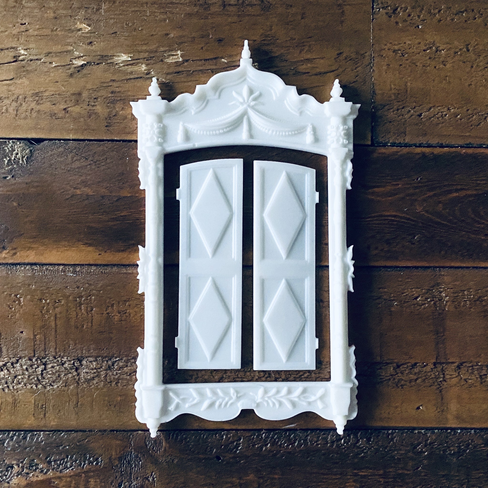 Three white resin castings of an ornate vintage window frame and 2 shutters created from LaBlanche's Vintage Window silicone mold are against a wood background.