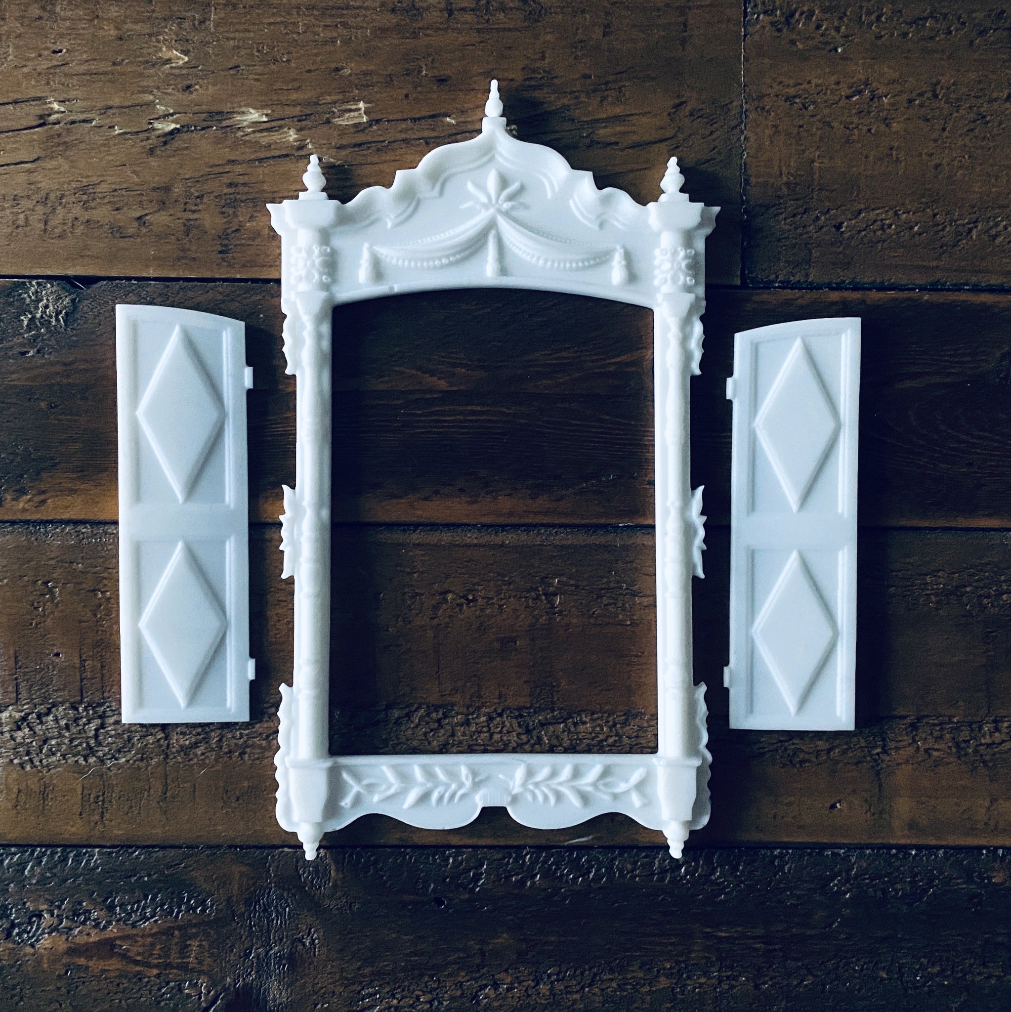 Three white resin castings of an ornate vintage window frame and 2 shutters created from LaBlanche's Vintage Window silicone mold are against a wood background.