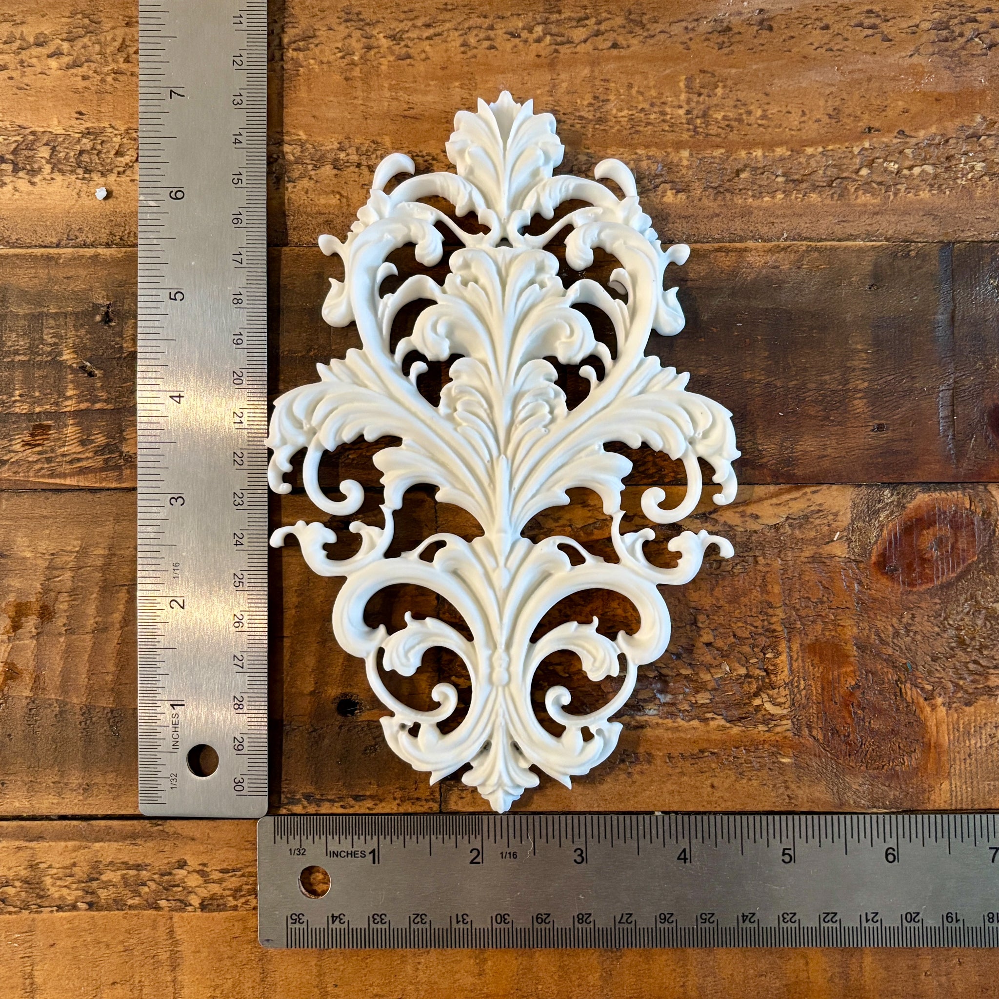 A white resin casting created from LaBlanche's Baroque Emblem silicone mold is against a dark wood background. Two silver rulers reading 7" height and 4.5" wide are to the left and under the casting.