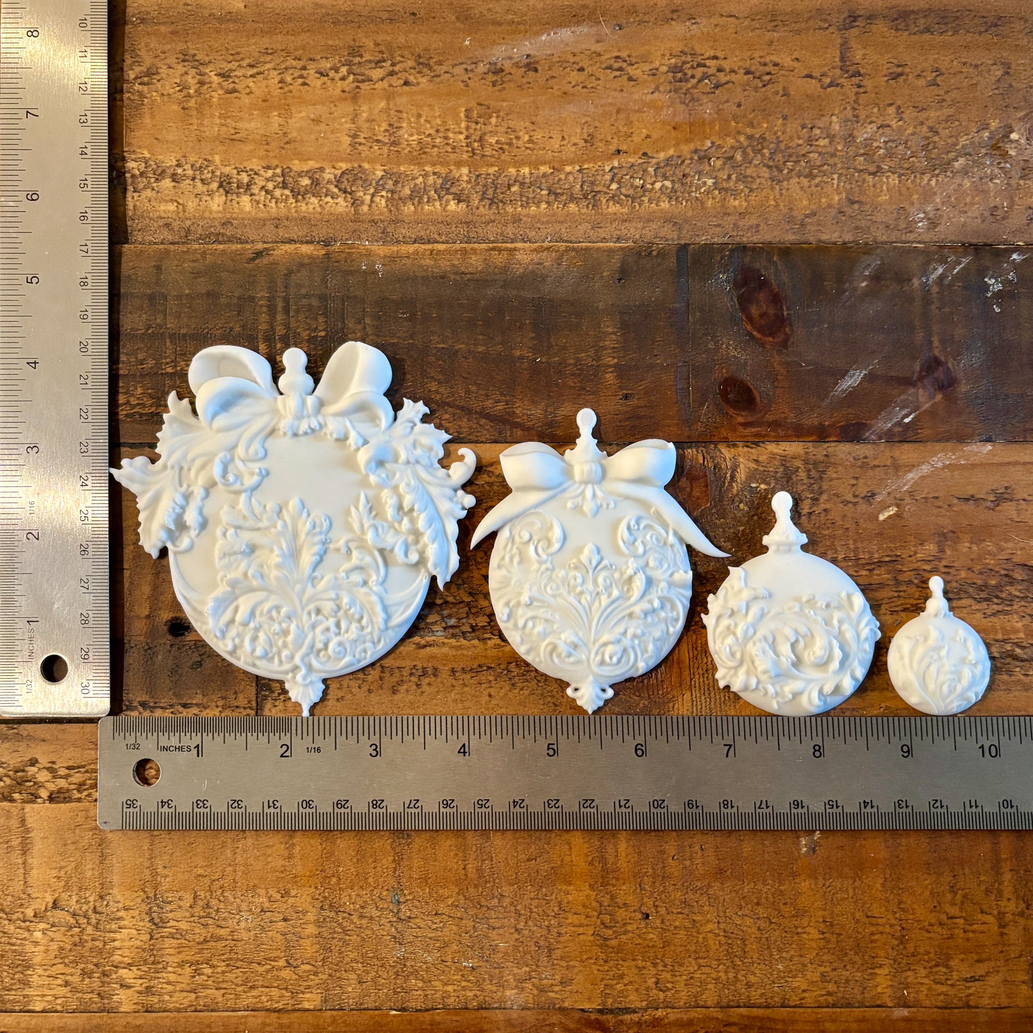 Four white resin castings of ornate Christmas ball ornaments are against a wood background. Two silver rulers to the left and under the castings read 4" height for the largest casting and a total of 10" length for all 4 castings.