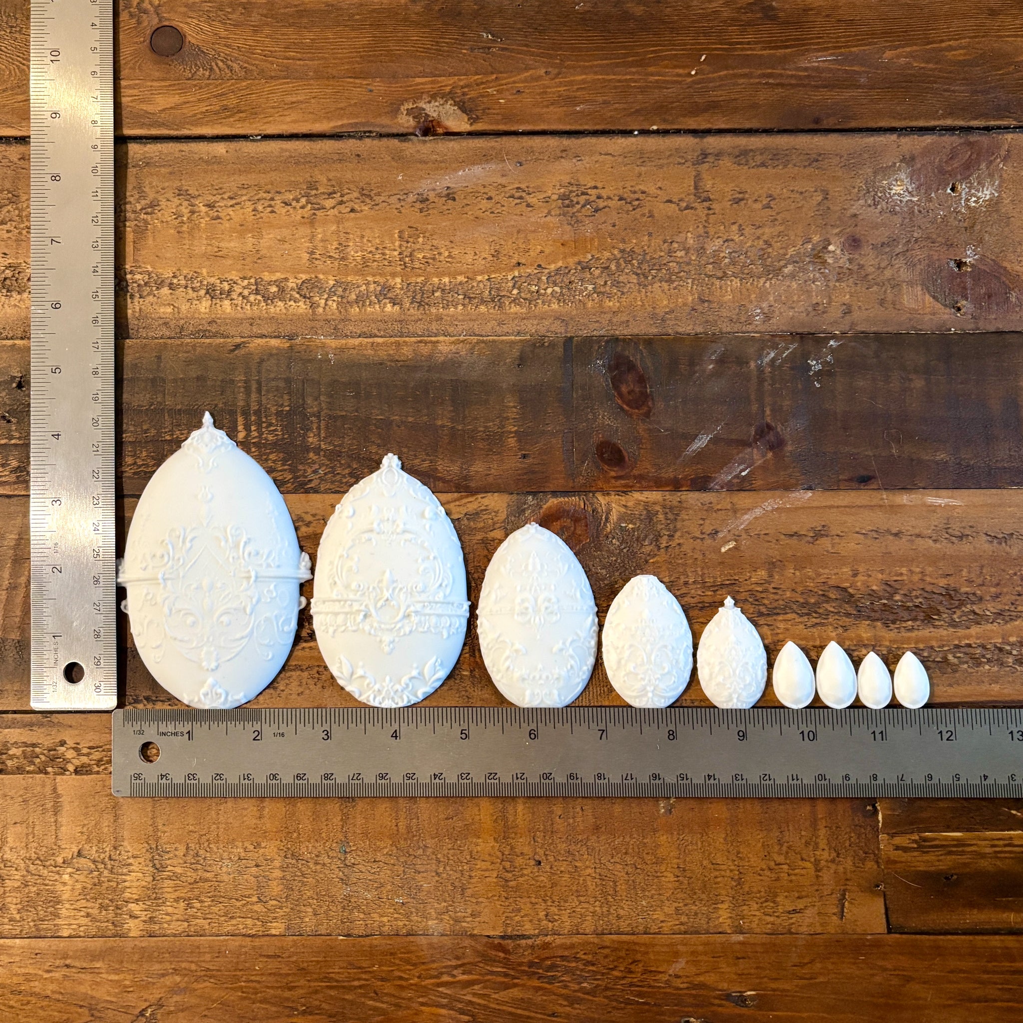 White silicone mold castings of 5 varying ornately detailed Easter eggs and 4 small teardrop pendants are against a wood background. Two silver rulers sit to the left and under the castings and read 4.25" height for the largest egg, and a total of 11.5" length for all the castings.