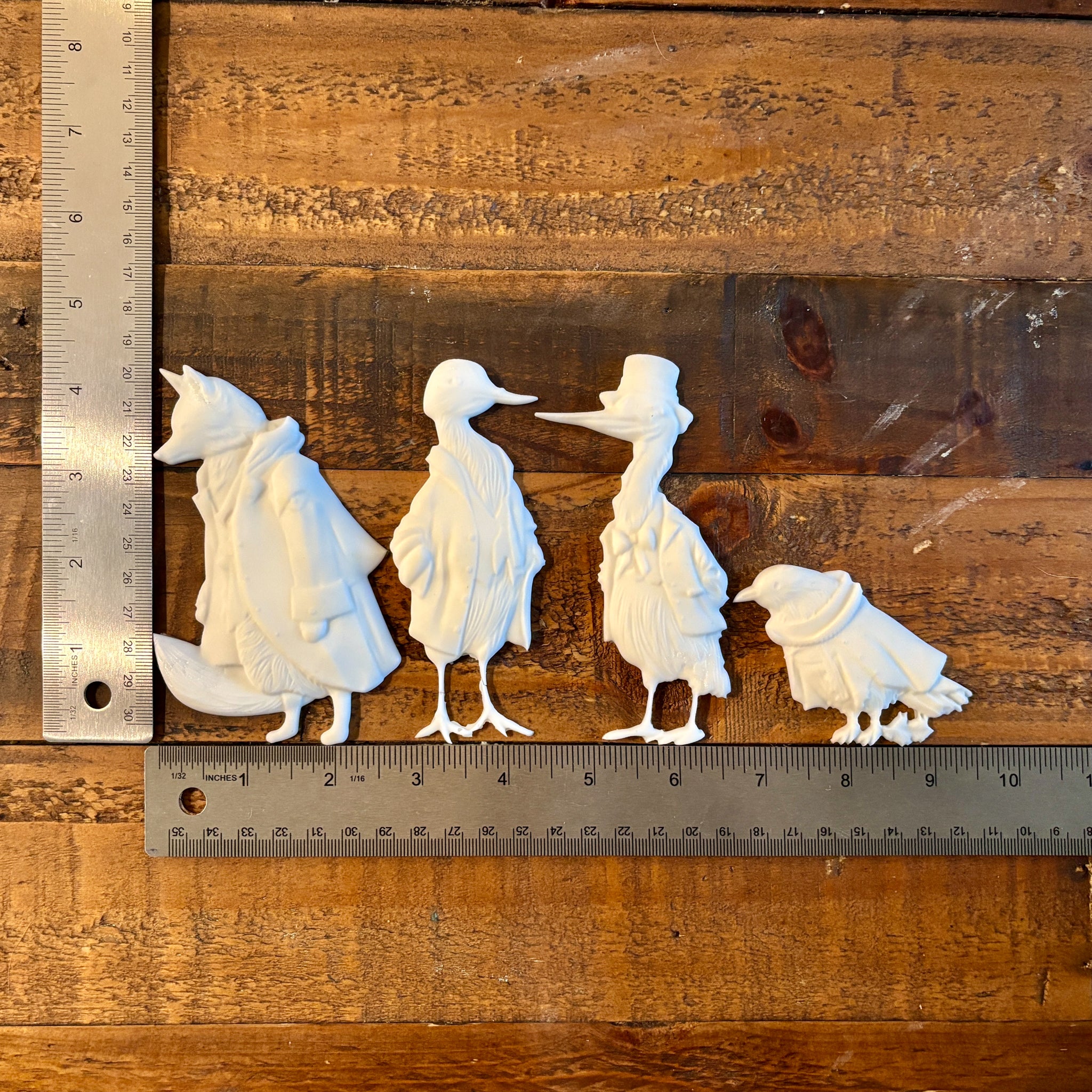 White resin castings of a fox and 3 birds all wearing vintage overcoats are against a wood background. Two silver rulers reading 4.5" height for the tallest castings and a total of 9.5" length for all 4 castings sit to the left and under the castings.