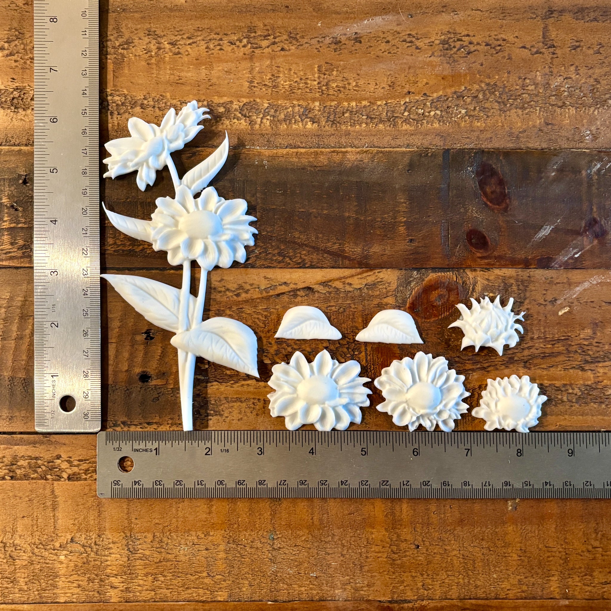 White resin castings of gerbera daisies are against a dark wood background. Two silver rulers reading 6.25" height for the tallest daisies and a total of 8.5" length for the rest of the castings sit to the left and under the castings.