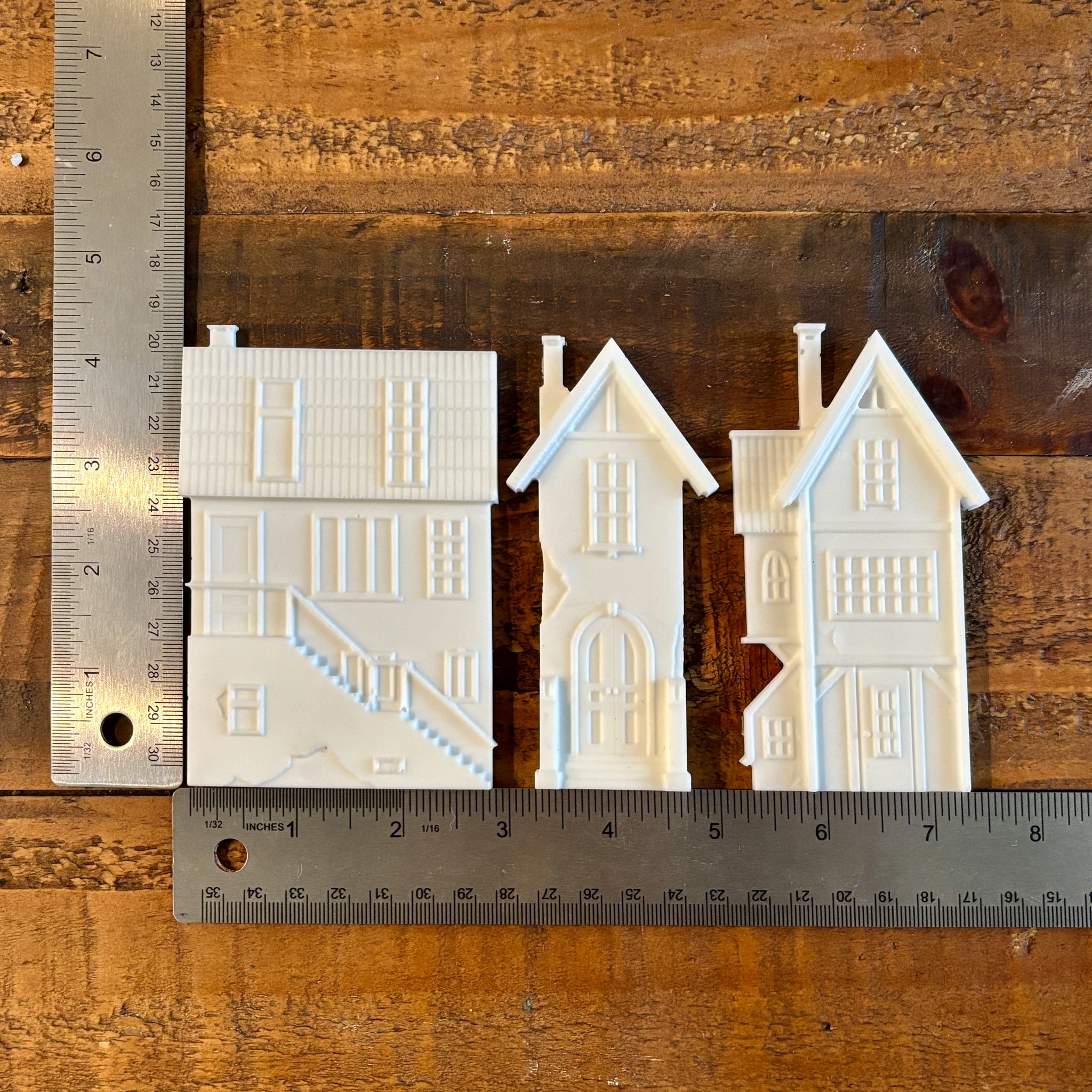 White resin castings of three 3-story houses are against a wood background. Two silver rulers sit to the left and under the castings; they read 4.25" height and 7.5" length with the houses slightly spaced apart.