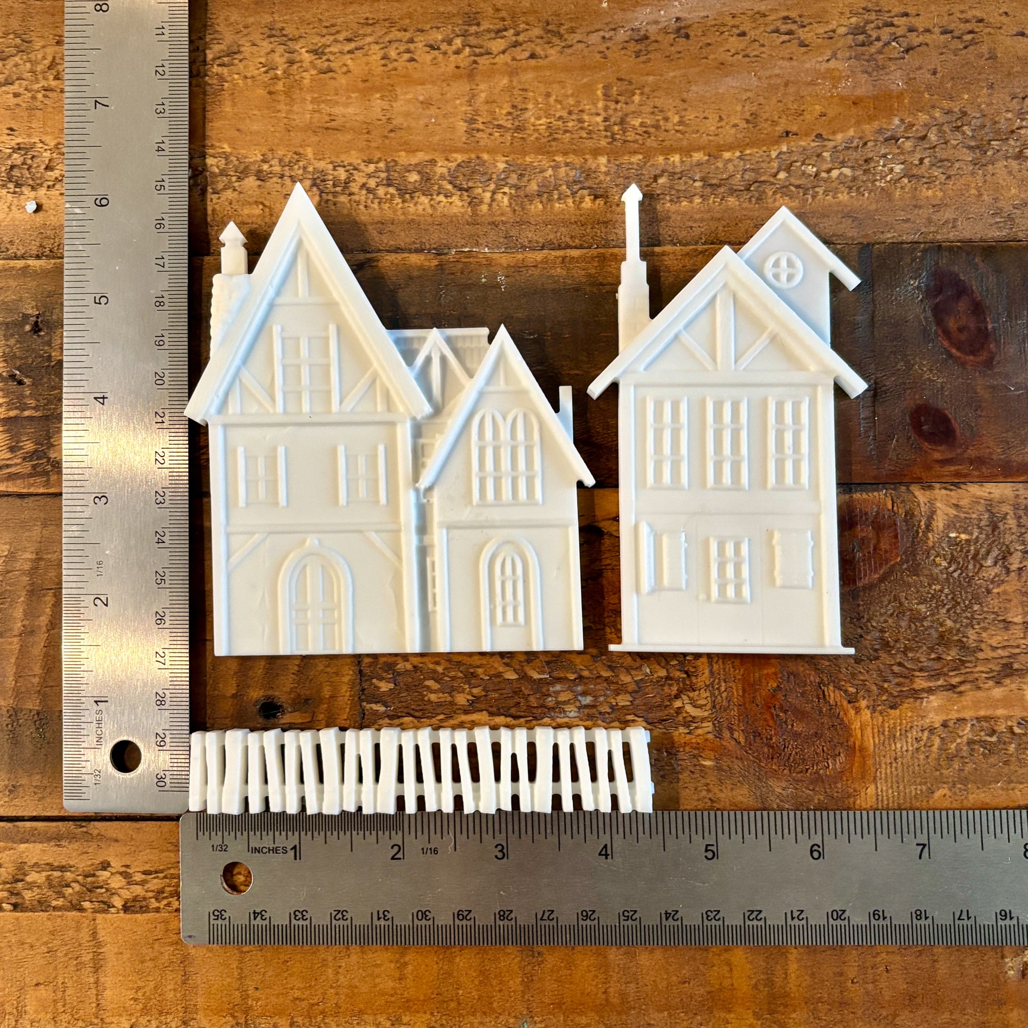 White resin castings of two 3-story houses and a picket fence are against a wood background. Two silver rulers sit to the left and under the castings; they read 6.25" height and 6.5" length with the houses slightly spaced apart and the fence under them.