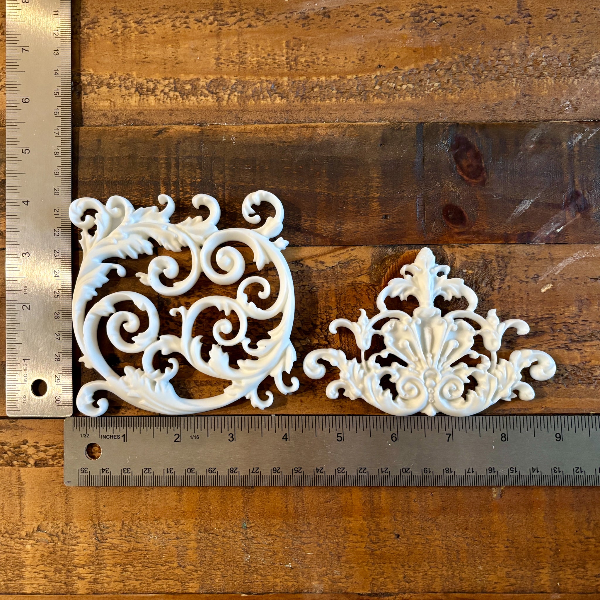 Two white resin castings of ornate scroll center accent pieces are against a wood background. Two silver rulers reading 4" height by 9" length sit to the left and under the castings.