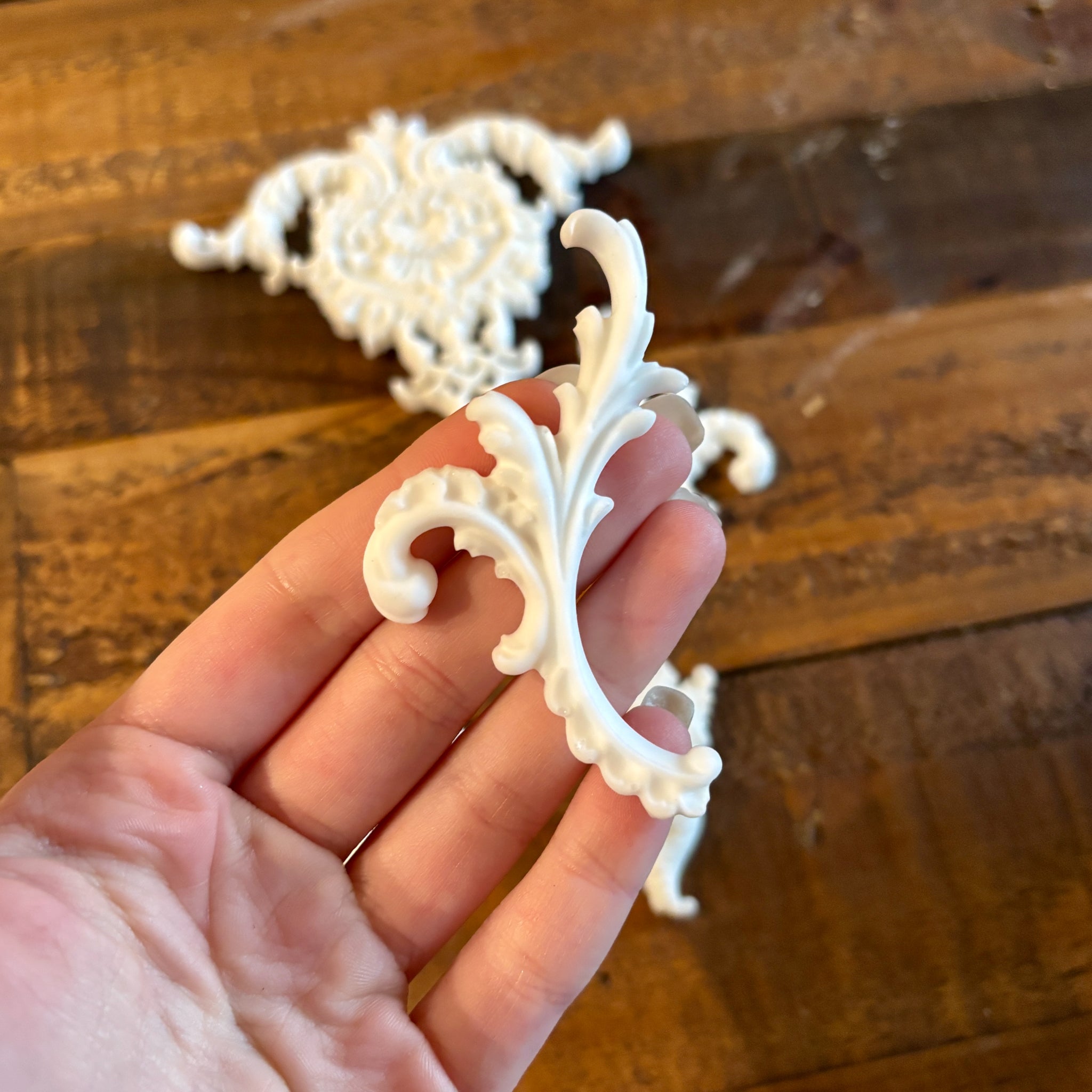 White resin castings of 4 small, and 1 large center ornate scroll accent pieces created using LaBlanche's Ornate Accents silicone mold are against a wood background. a hand is shown holding one of the small castings.