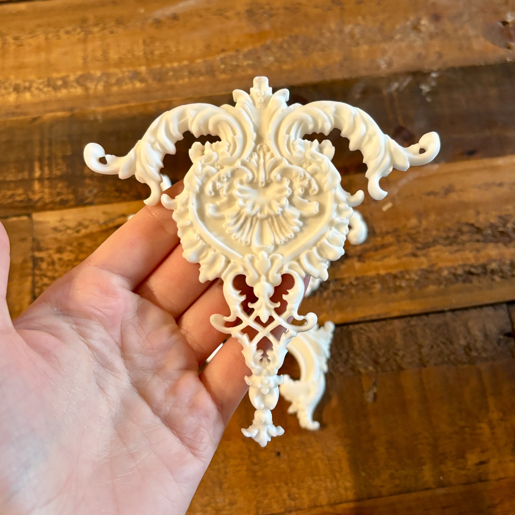 A hand is shown holding a large ornate center accent white resin casting against a wood background.