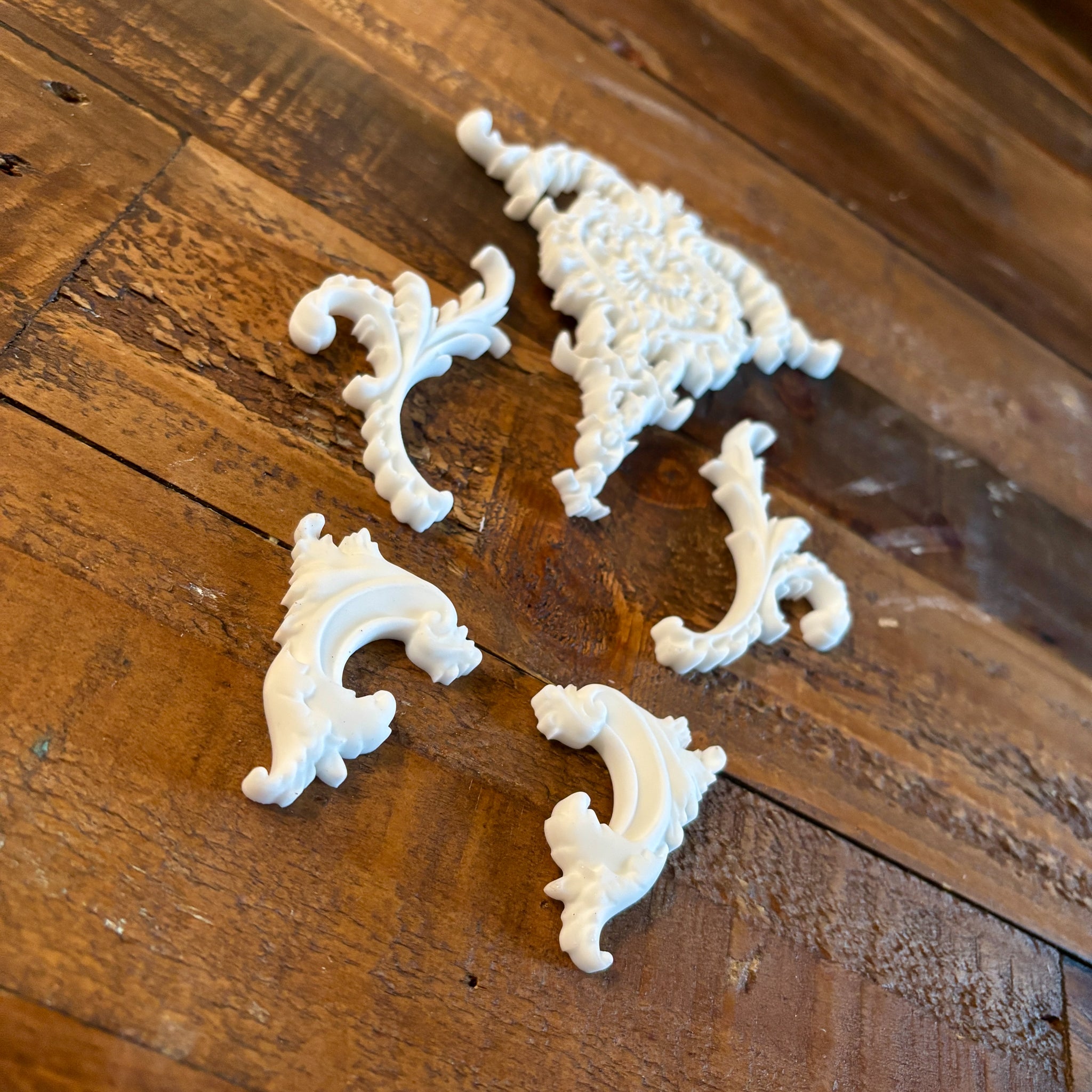 An angled view of white resin castings of 4 small, and 1 large center ornate scroll accent pieces created using LaBlanche's Ornate Accents silicone mold are against a wood background.