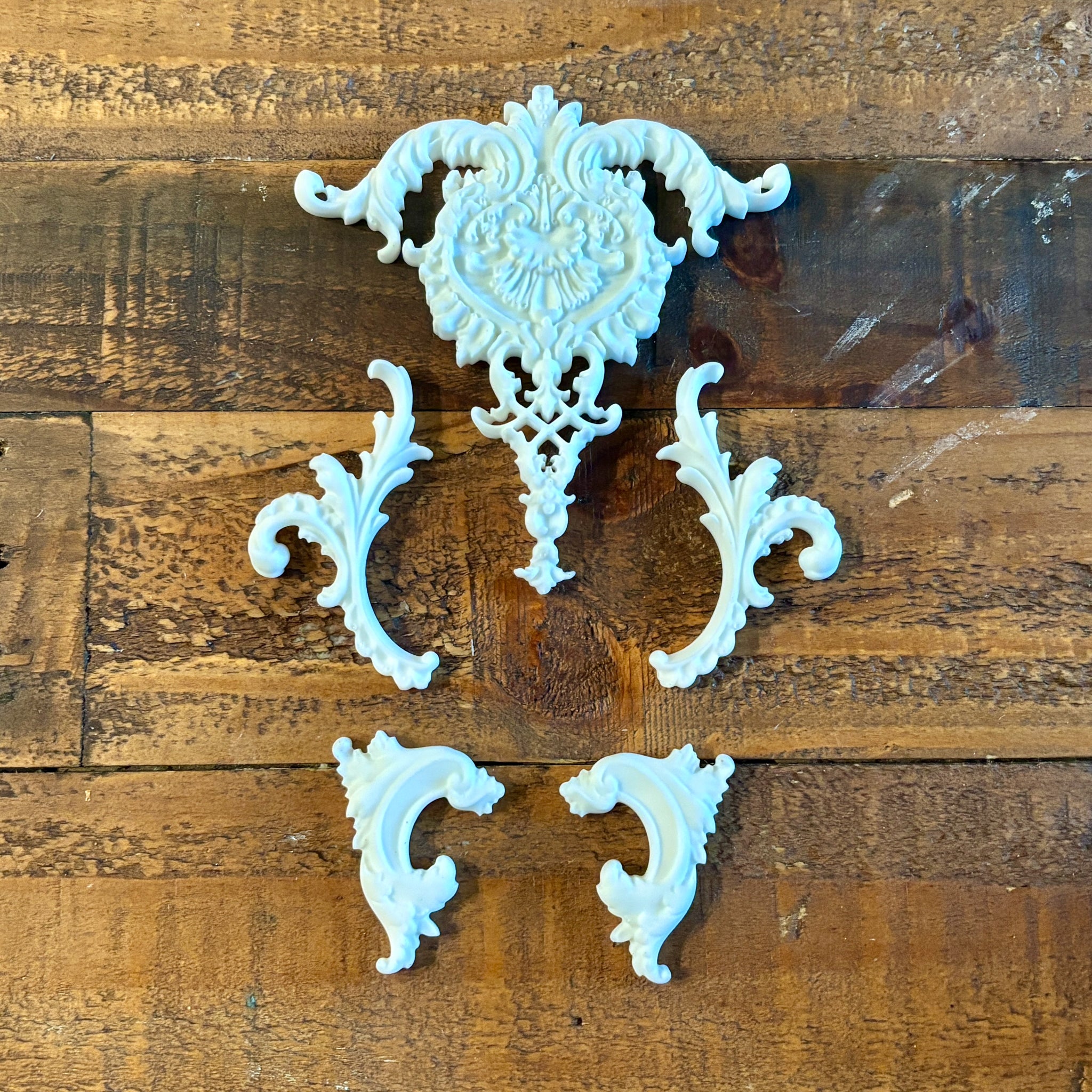 White resin castings of 4 small, and 1 large center ornate scroll accent pieces created using LaBlanche's Ornate Accents silicone mold are against a wood background.