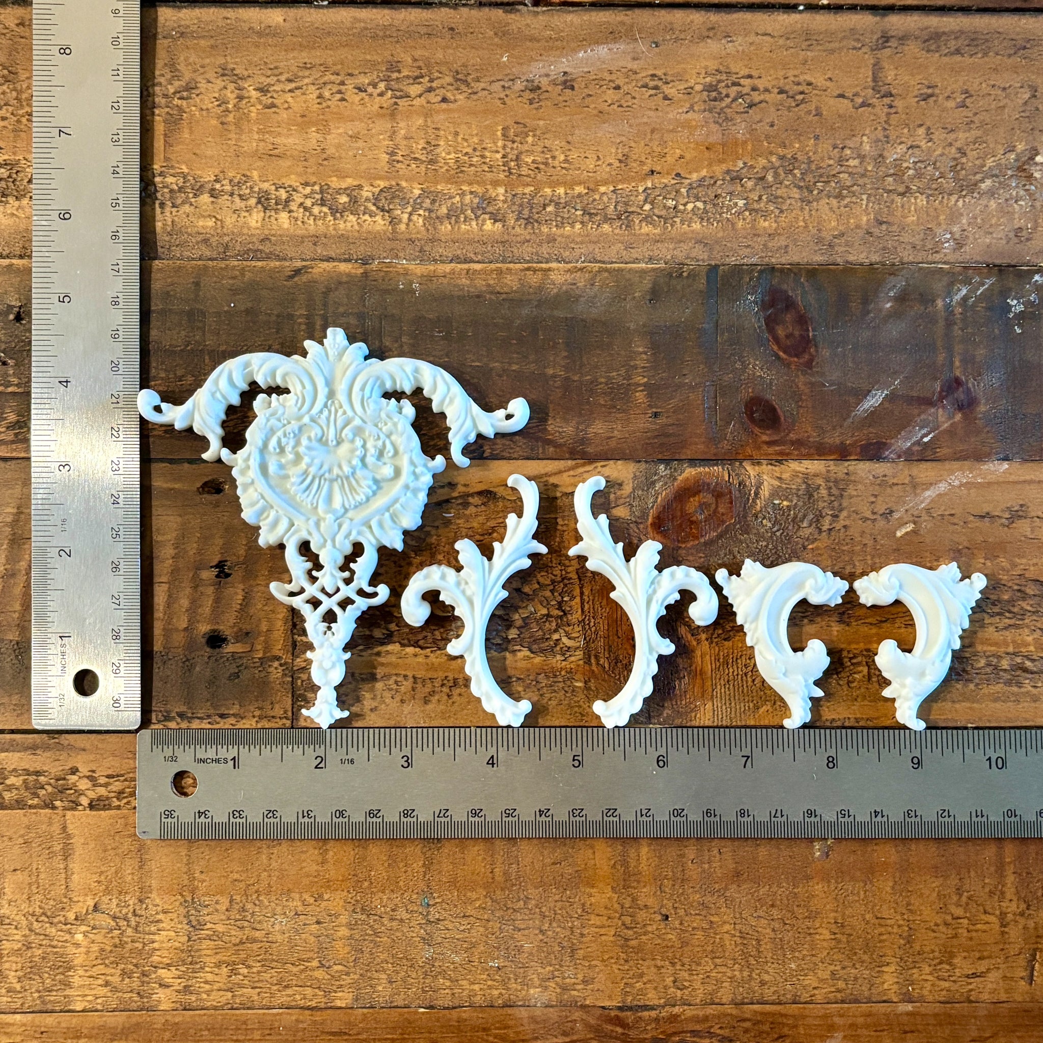 Four white resin castings created from LaBlanche's Ornate Accents silicone mold are lined up on a wood background. Two silver rulers reading 4.5" height by 10" length sit to the left and under the castings.