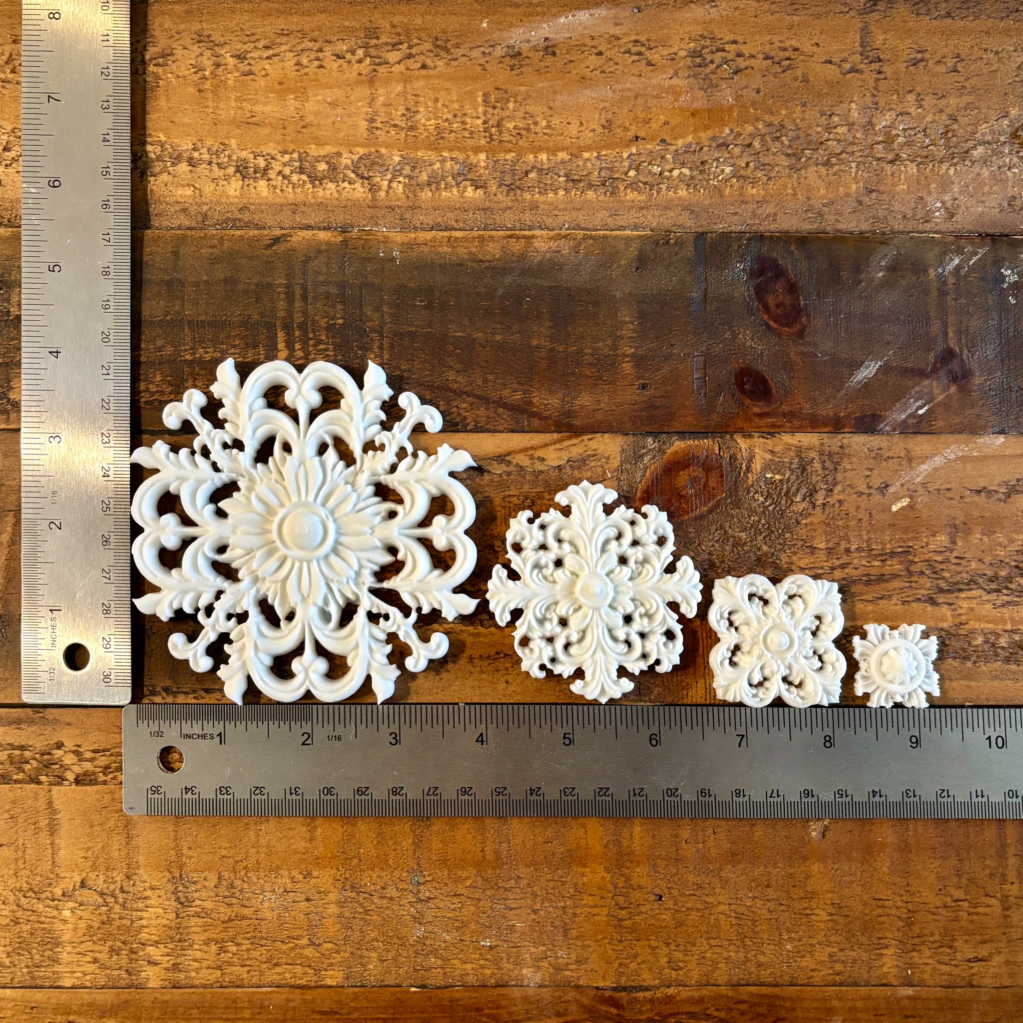 White resin castings of 4 varying size round ornate flourish medallions are against a dark wood background. Two silver rulers reading 4" height for the largest casting and 9" length for all 4 castings next to each other sit to the left and under the castings.