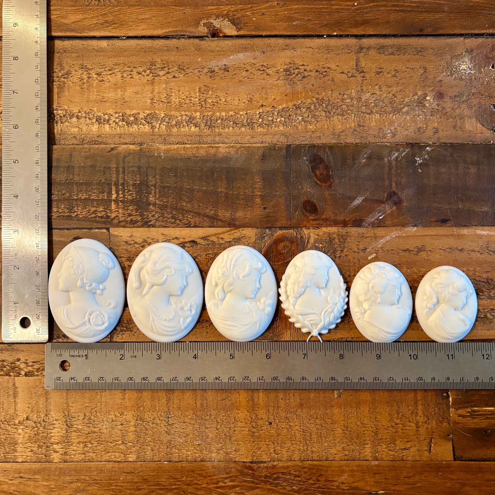 Six white resin castings of cameo pendants created from LaBlanche's Vintage Cameo silicone mold are lined up on a wood background. Two silver rulers reading 2.75" height by 11.5" length sit to the left and under the castings.