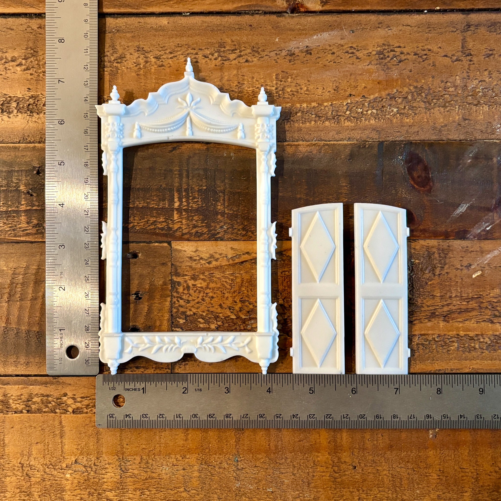 White resin castings of a vintage window frame and 2 shutters created from LaBlanche's Vintage Window silicone mold are lined up on a wood background. Two silver rulers reading 7.75" height by 7.25" length sit to the left and under the castings.