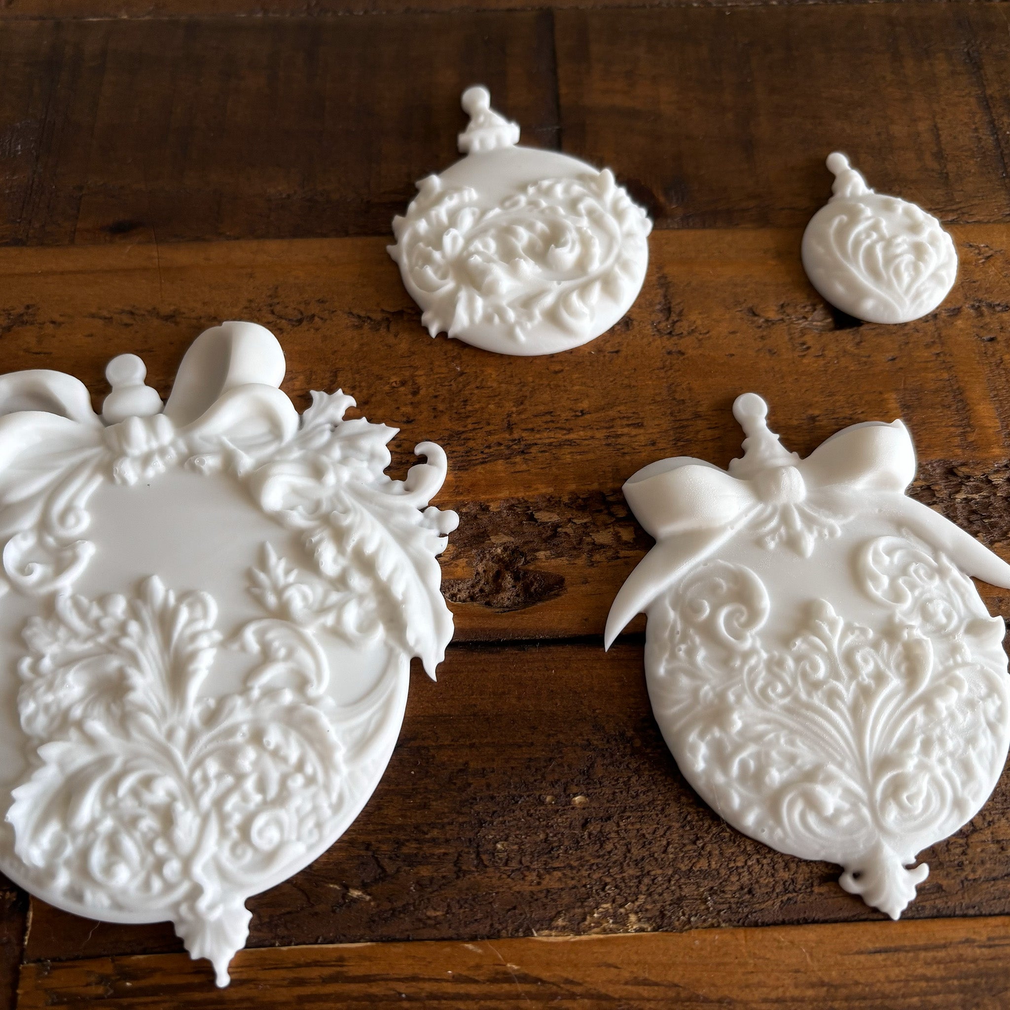 Four white resin castings of ornate Christmas ball ornaments in varying sizes created from LaBlanche's Christmas Balls silicone mold are against a dark wood background.