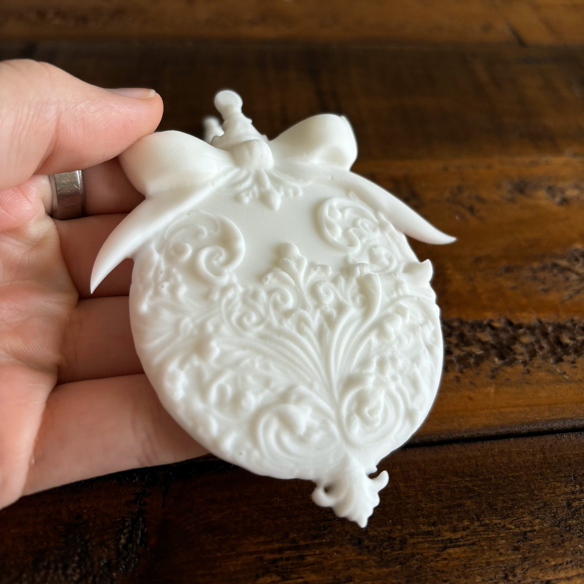 A hand is shown holding a white resin casting of a large Christmas ball ornament against a dark wood background.