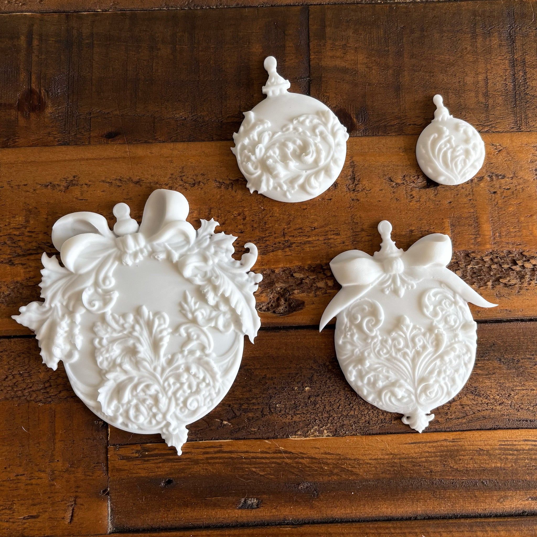 Four white resin castings of ornate Christmas ball ornaments in varying sizes created from LaBlanche's Christmas Balls silicone mold are against a dark wood background.