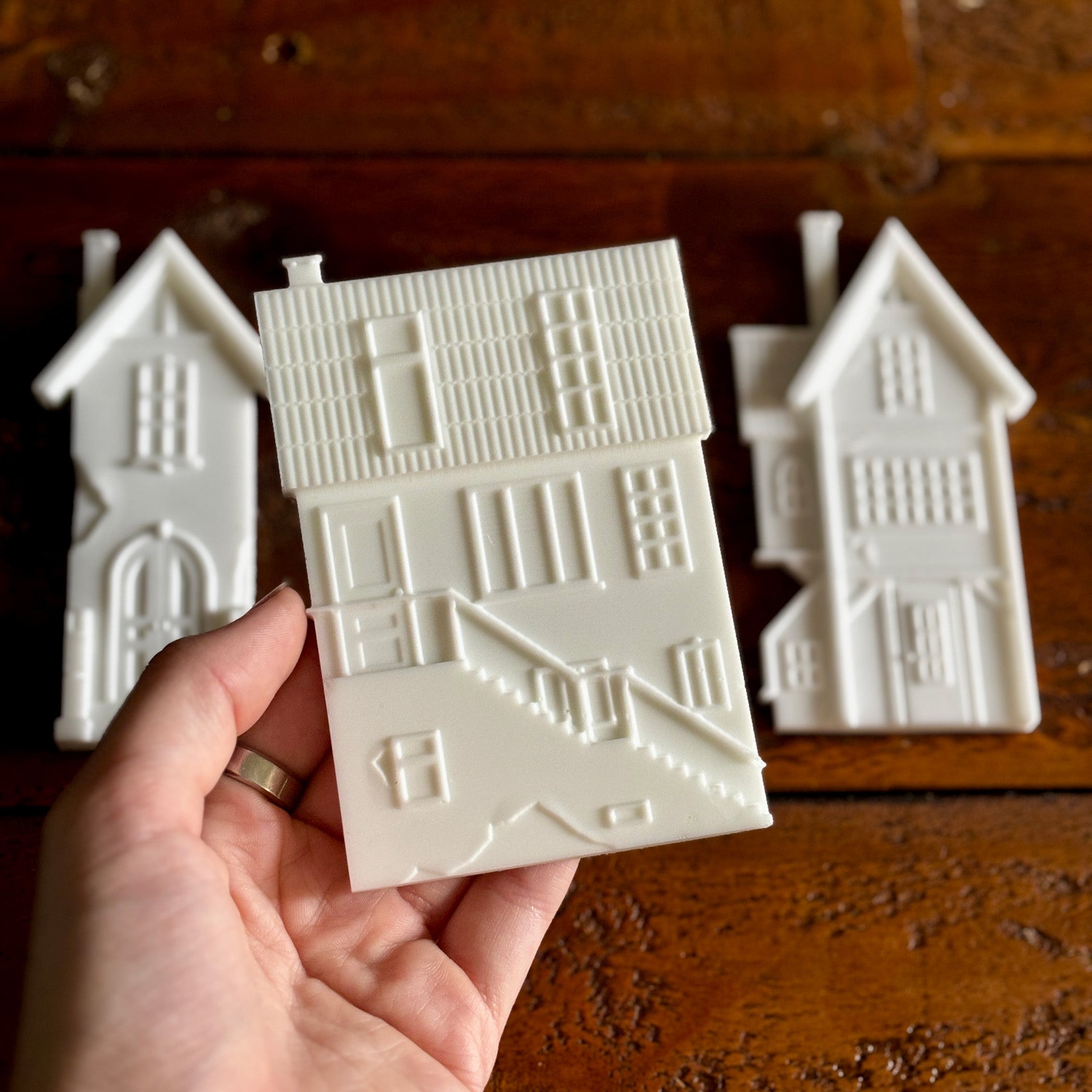 White resin silicone mold castings of 3 houses are against a dark wood background. A hand is shown holding one of the houses.