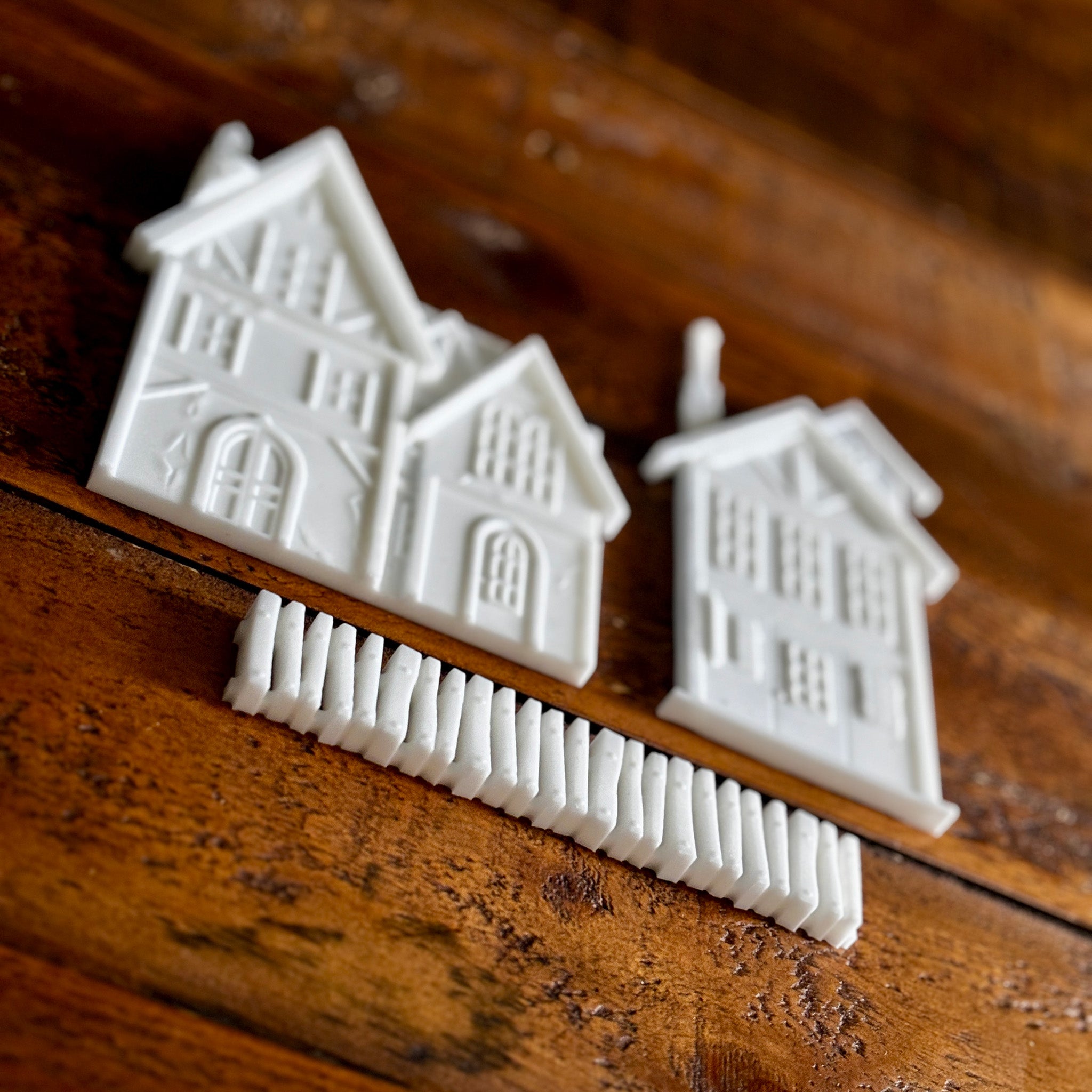 White resin silicone mold castings of 2 houses and a picket fence are against a dark wood background.