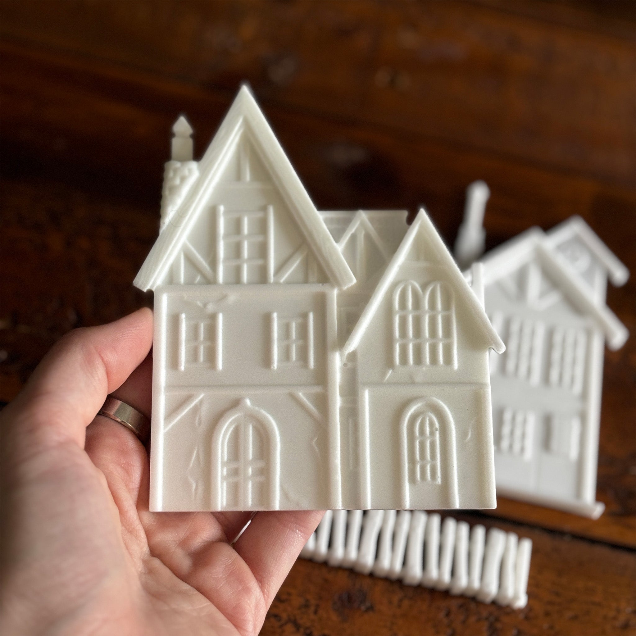 White resin silicone mold castings of 2 houses and a picket fence are against a dark wood background. A hand is shown holding one of the houses.