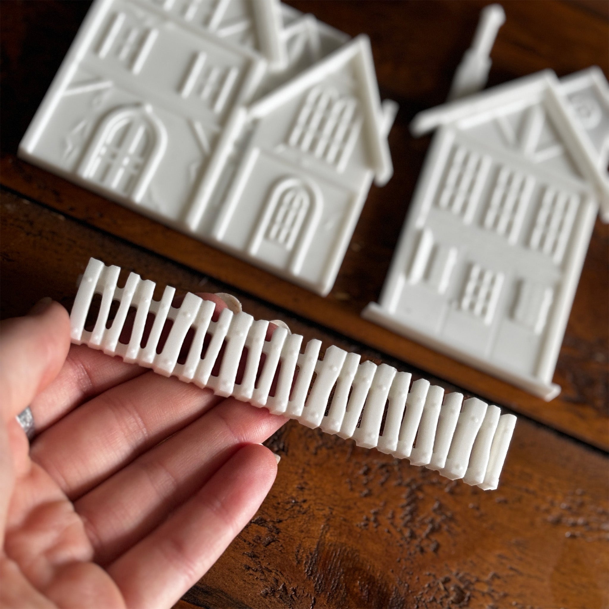 White resin silicone mold castings of 2 houses and a picket fence are against a dark wood background. A hand is shown holding the picket fence.