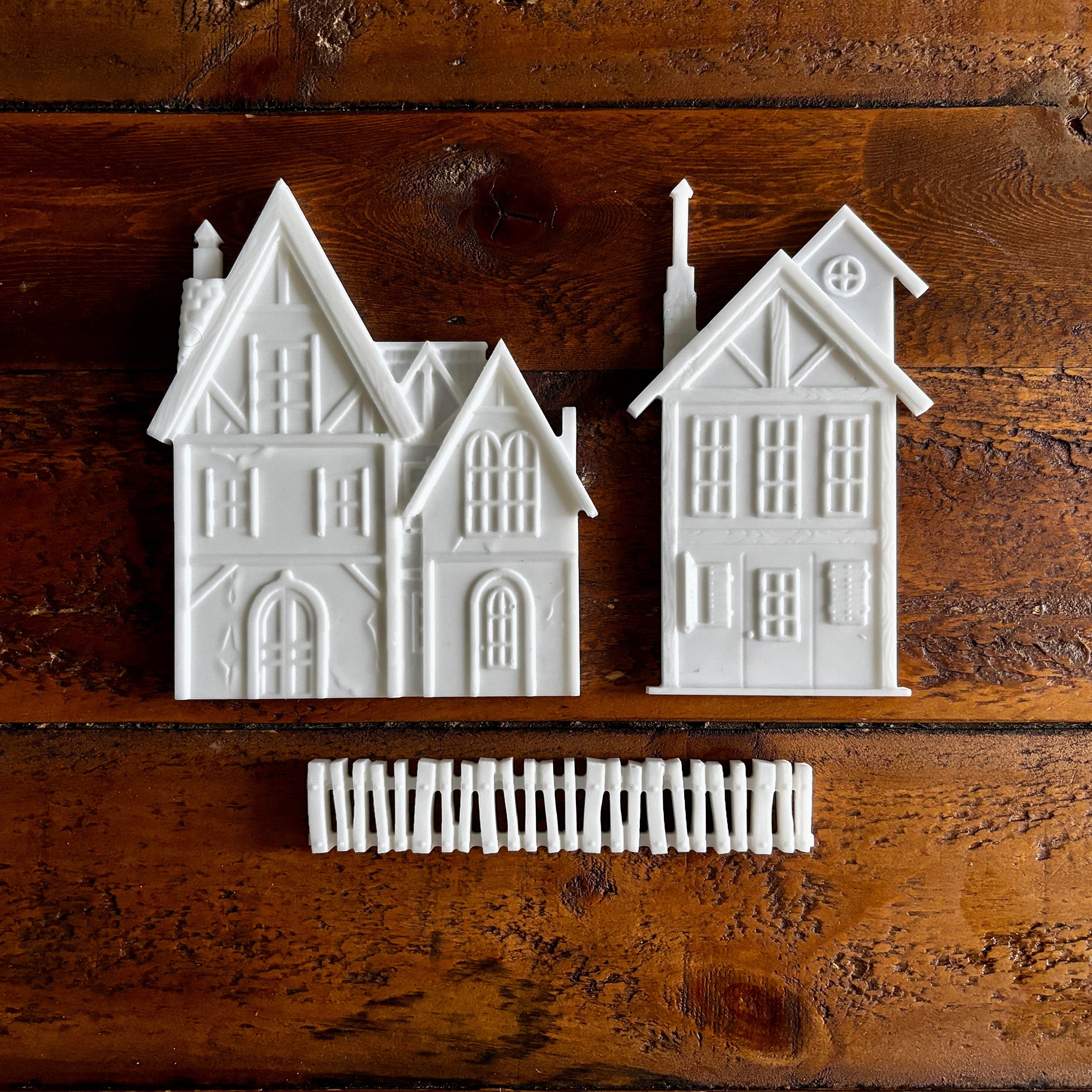 White resin silicone mold castings of 2 houses and a picket fence are against a dark wood background.