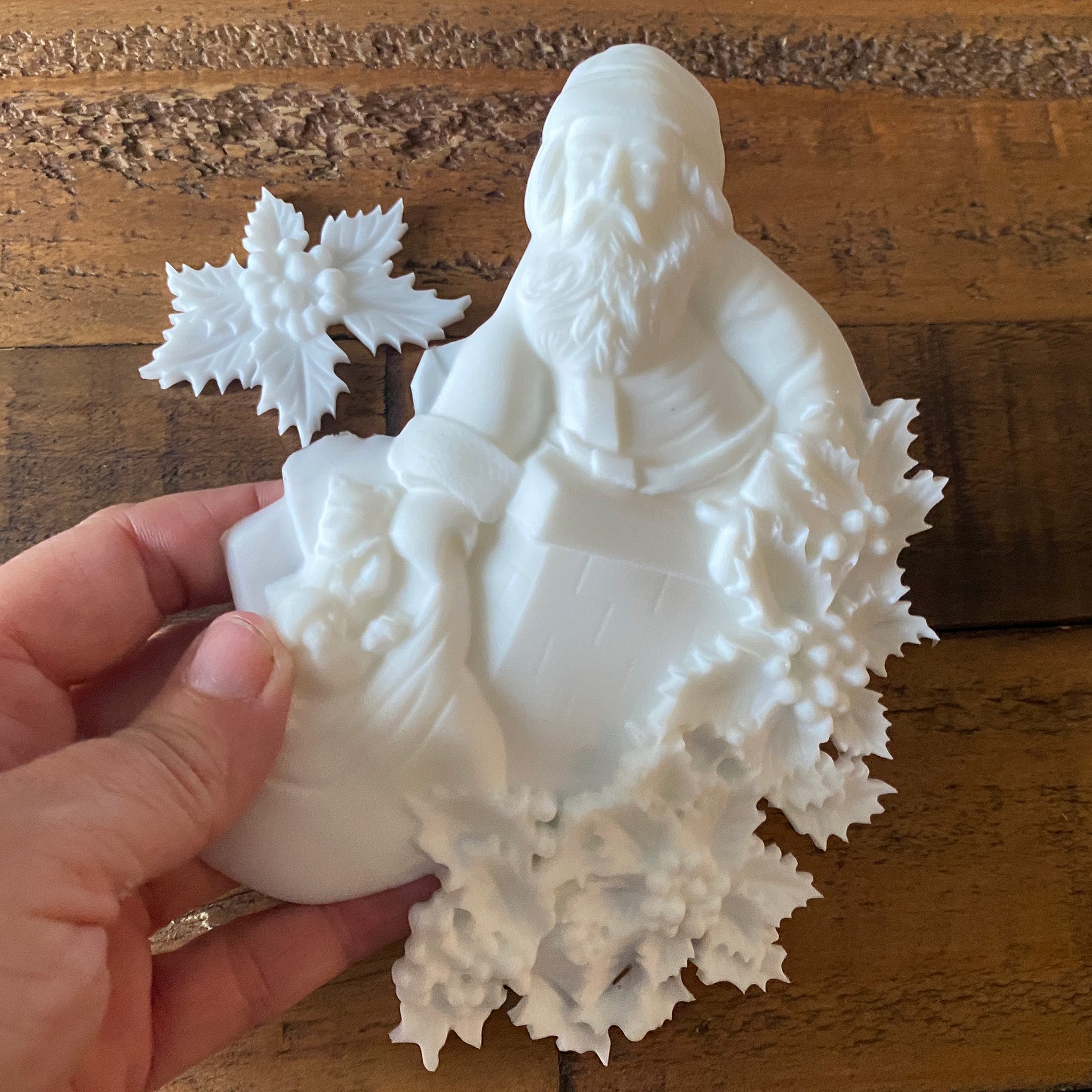 Two white resin castings made from LaBlanche's St Nikolaus silicone mold of Santa Claus with a bag of gifts and holly garland and a single holly with berries are against a dark wood background. A hand is shown holding the Santa casting.