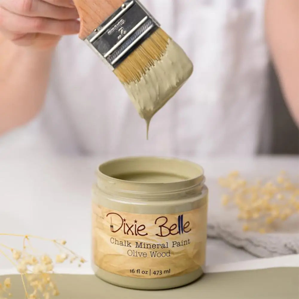 A paint brush with dripping paint is shown above an open container of Dixie Belle's Olive Wood Chalk Mineral Paint.