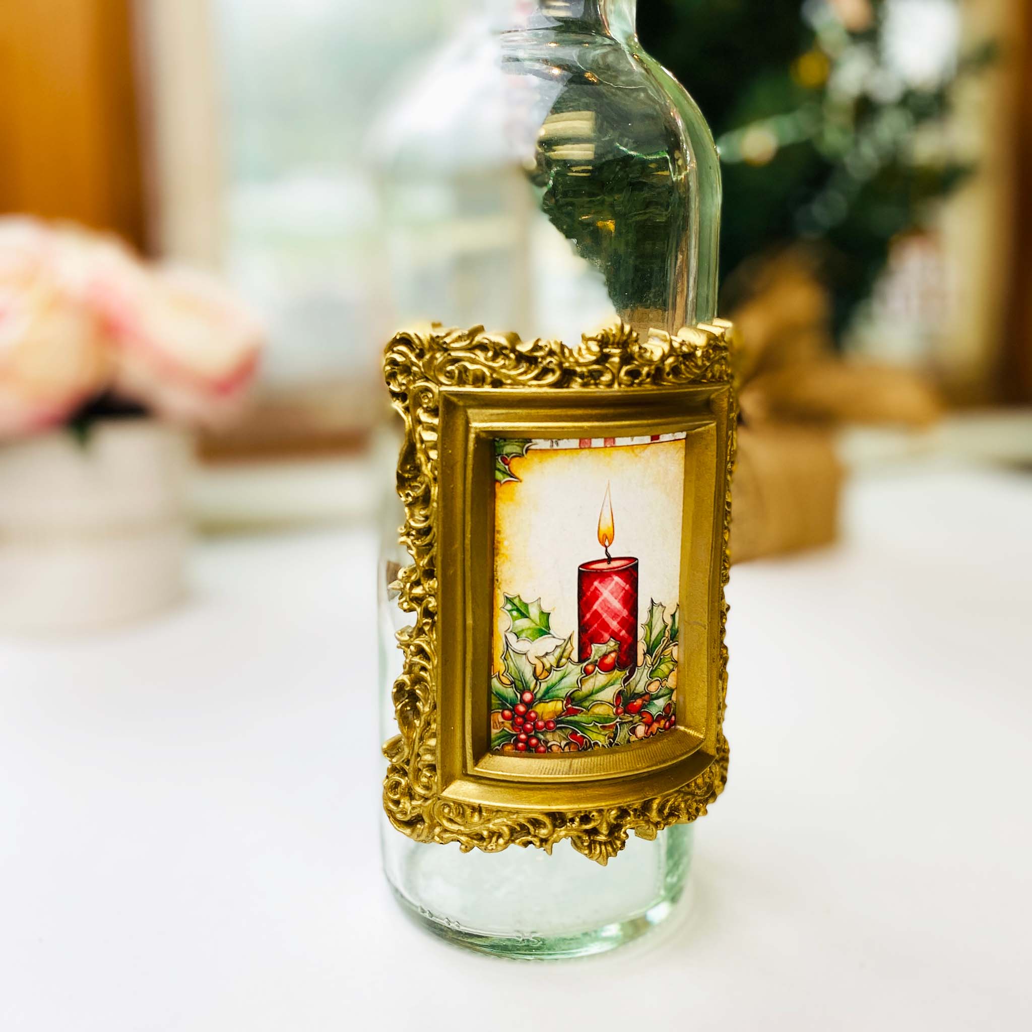 A clear glass bottle has a small silicone casting of an ornate picture frame is covered with Pentart's metallic gold wax paste. A small photo of a holiday candle with holly berries is inside the frame.