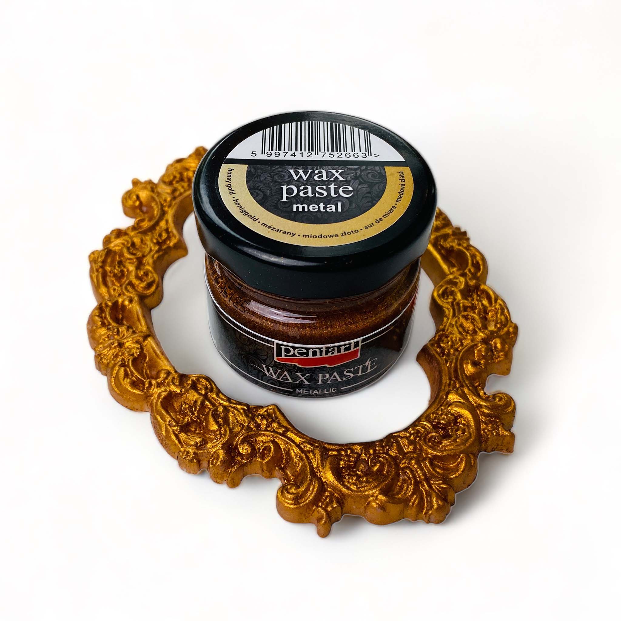 An open jar of a 20ml/0.68 ounce jar of metallic Honey Gold Wax Paste by Pentart is against a white background. An ornate frame covered with Honey Gold Pentart wax paste surrounds the jar.