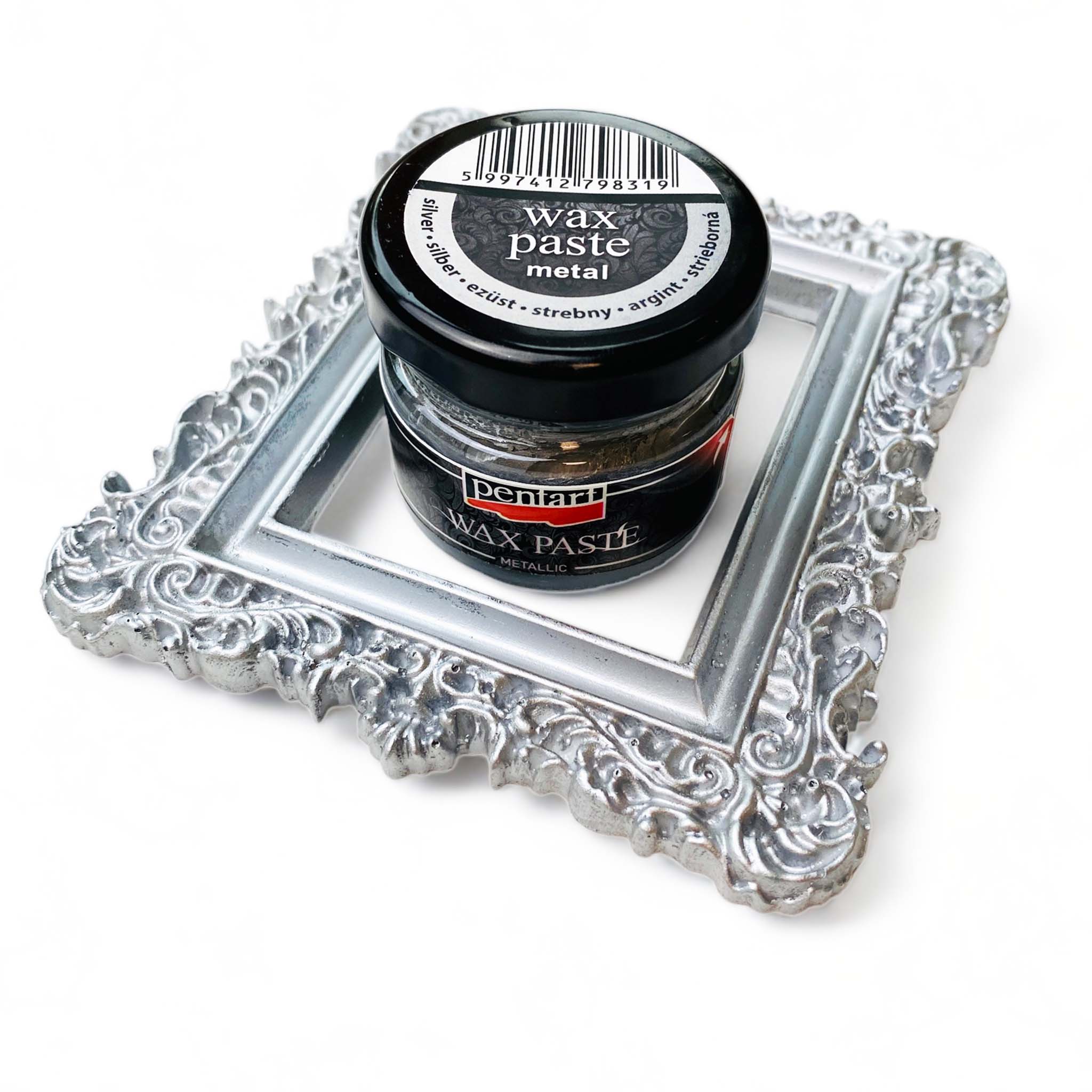 A jar of a 0.68 ounce jar of metallic silver wax paste by Pentart is sitting inside a silicone casting of an ornate picture frame covered with the wax. Both are on a white background.