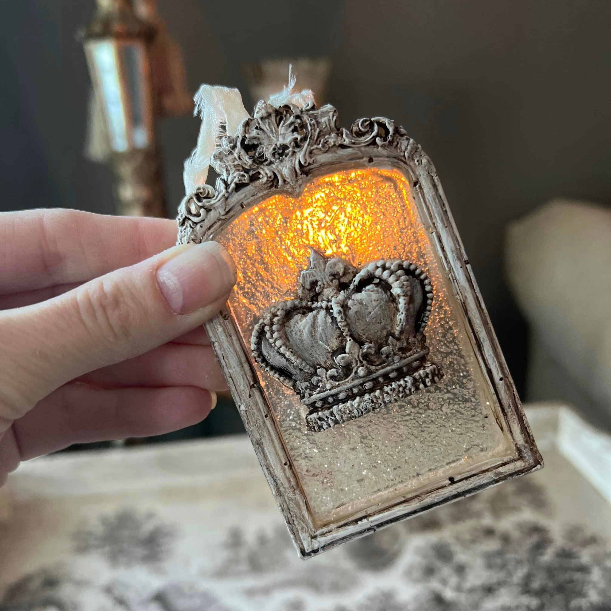 A small ornate silver colored frame featuring a clear resin center with a silver crown on it created from a silicone mold is being held in front of a light source to showcase the clear center.