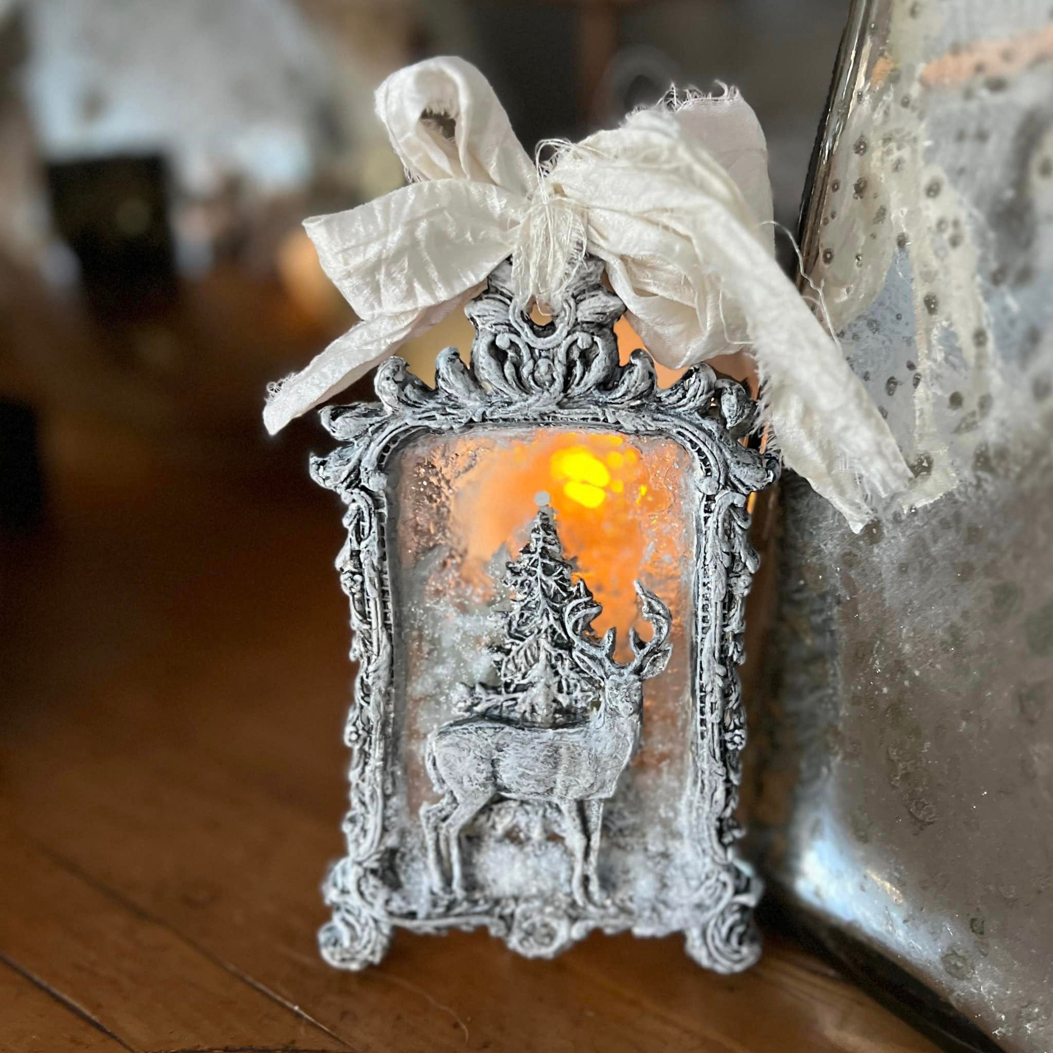 A small ornate silver colored frame featuring a clear resin center with a silver colored reindeer in front of a tree on it is set up in front of a small light course to showcase the clear resin.