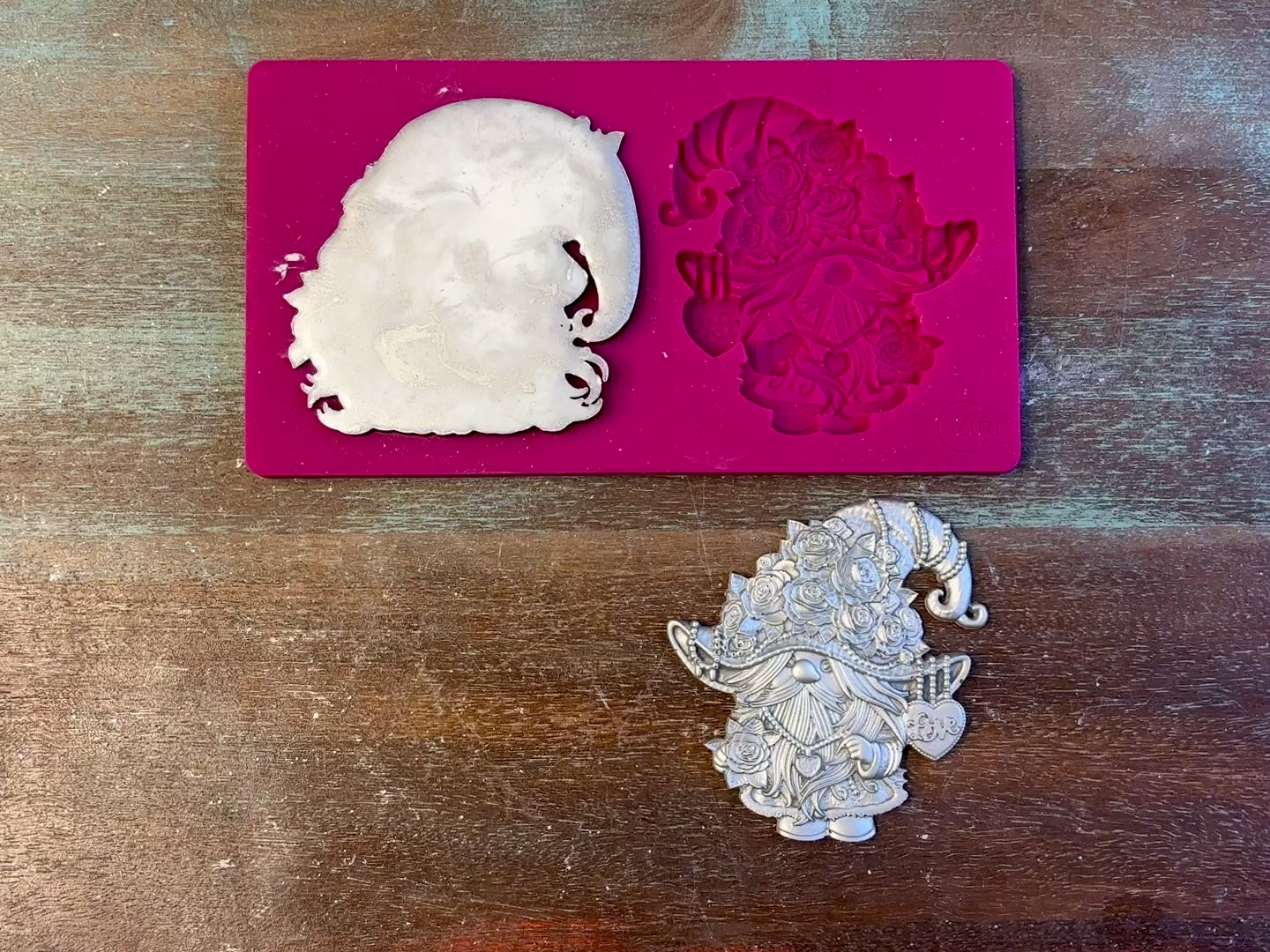 A 12 second video of Zuri Design's Valentine's Gnomes Set 2 silicone mold and silver colored castings are against a wood background. A hand is shown holding one of the castings.