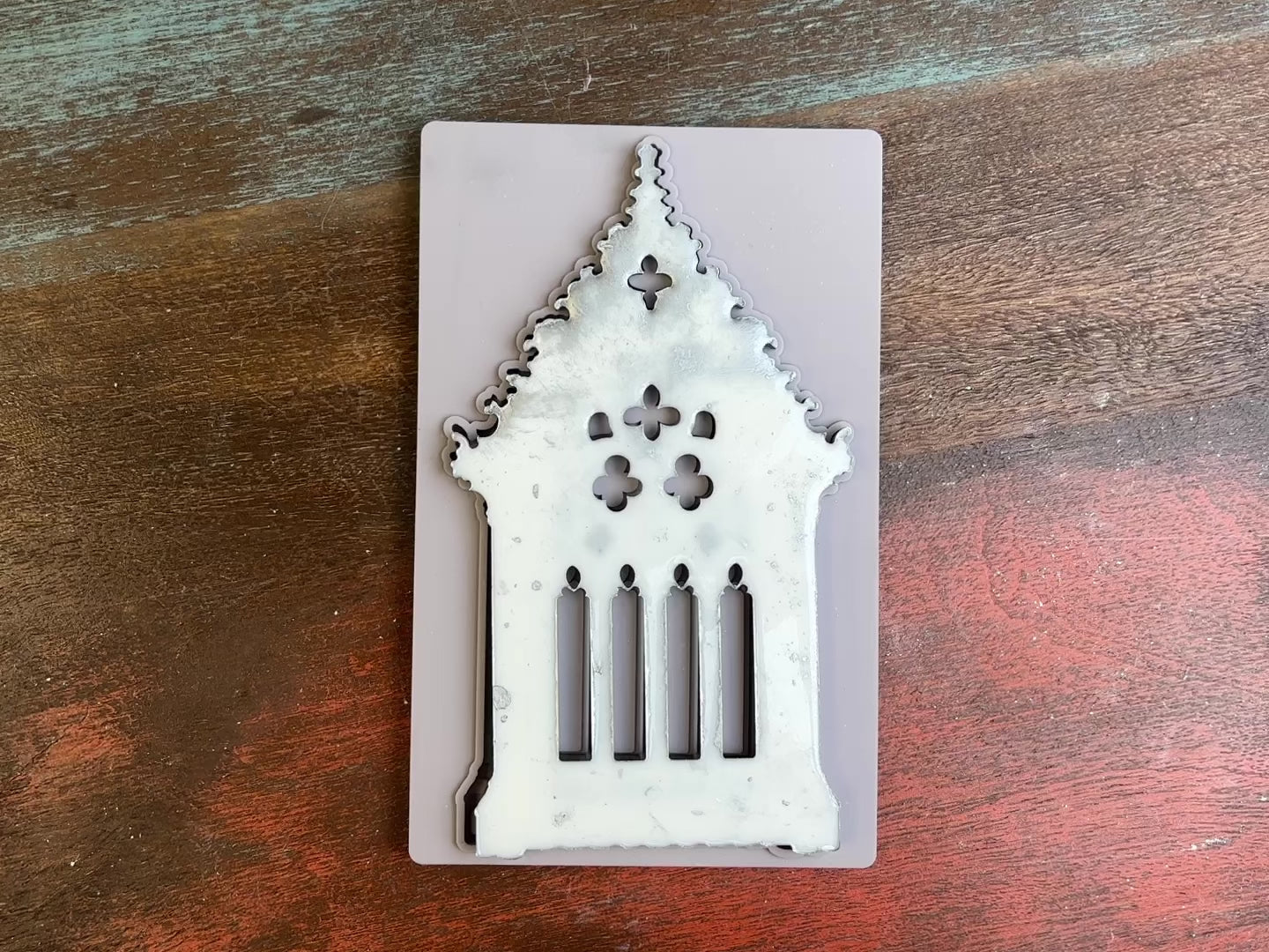 A 15 second video of a light grey silicone mold and silver colored casting of a gothic window are against a dark wood background. A hand is shown picking up and flipping over the casting to show the details.