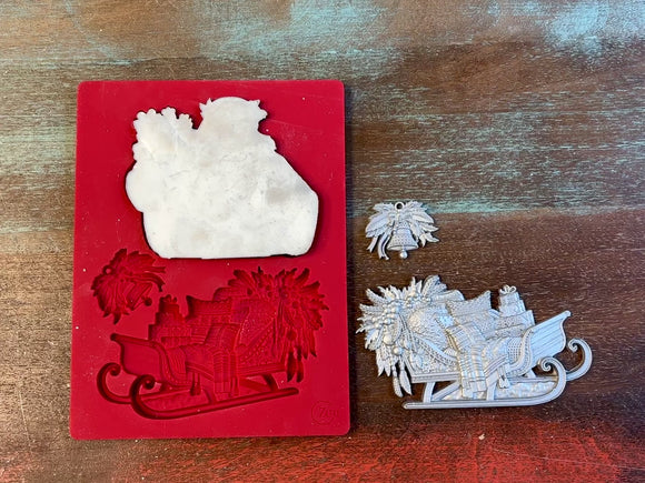 A 12 second video of Zuri Design's Bohemian Santa and Sled silicone mold and silver colored castings are against a wood background. A hand is shown holding the Santa casting.
