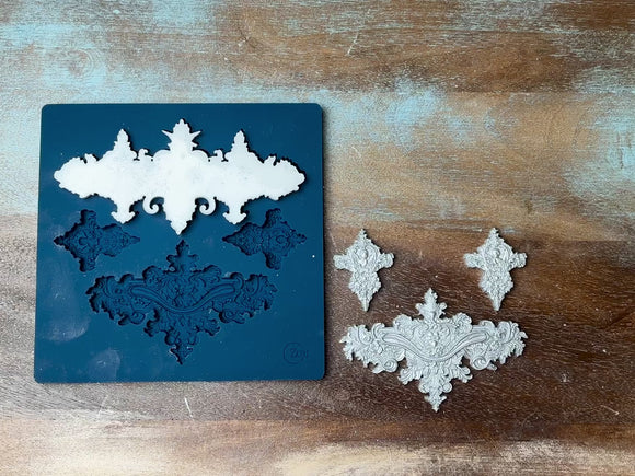 A 13 second video shows a close-up of Zuri Design's Baroque Ornaments Set 1 silicone mold and silver colored castings against a dark wood background. A hand is shown picking up one of the larger castings.