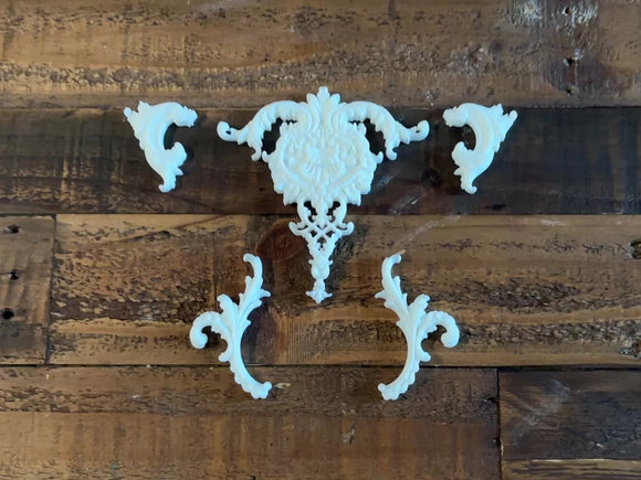 A 12 second video shows a close-up of 4 small, and 1 large ornate accent white resin castings created from LaBlanche's Ornate Accents silicone mold against a wood background. A hand is shown picking up one of the small castings.