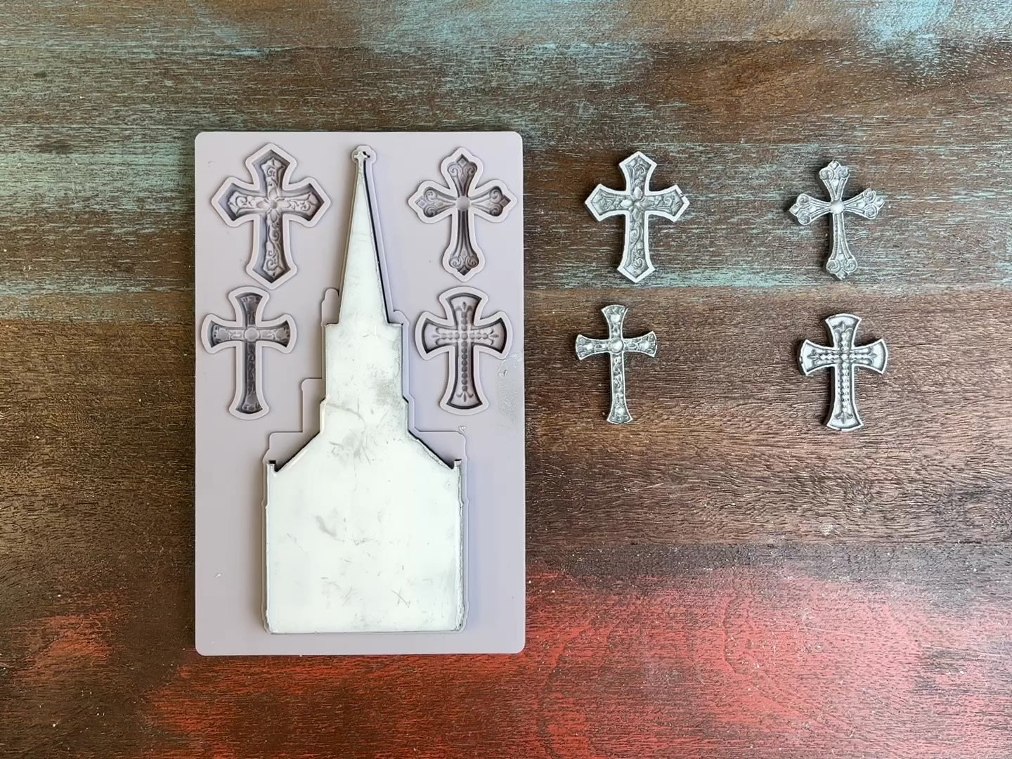 A 14 second video of a light grey silicone mold and silver colored castings of a church and 4 small ornate crosses are against a dark wood background. A hand is shown picking up and flipping over the church casting to show the details.