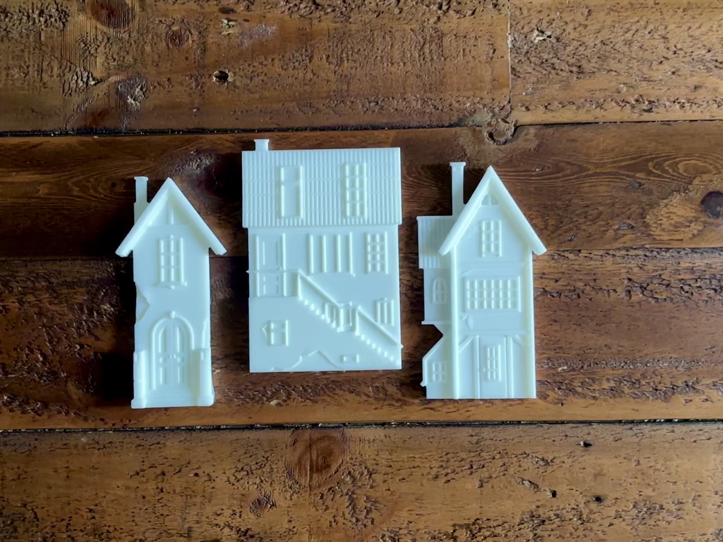 A 14 second video shows a close-up of 3 white resin silicone mold castings of houses against a dark wood background. A hand is shown picking up one of the houses.